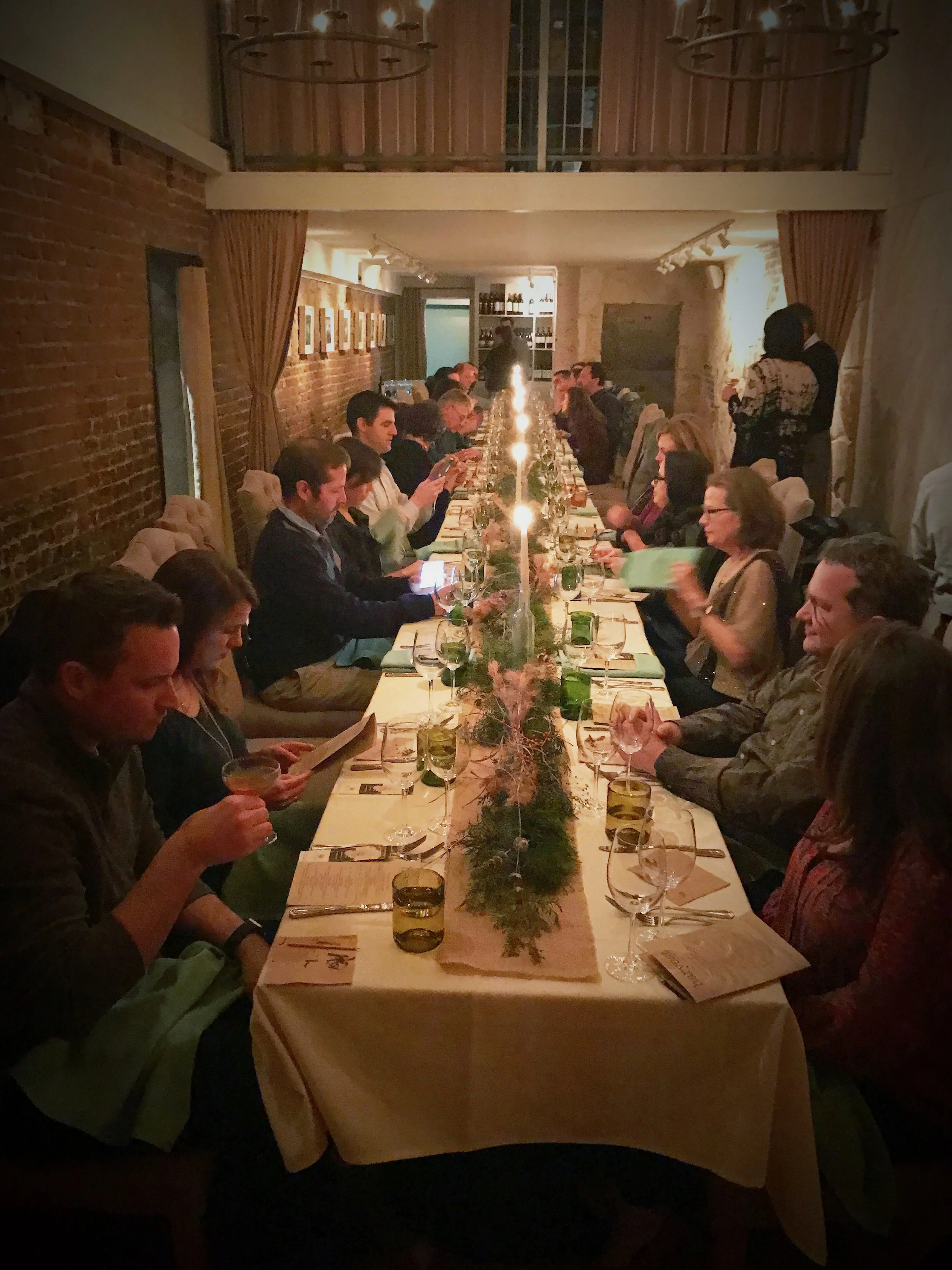 A long dinner table with many people seated on both sides, decorated with a green and brown centerpiece, in a warmly lit room with brick walls and framed pictures.