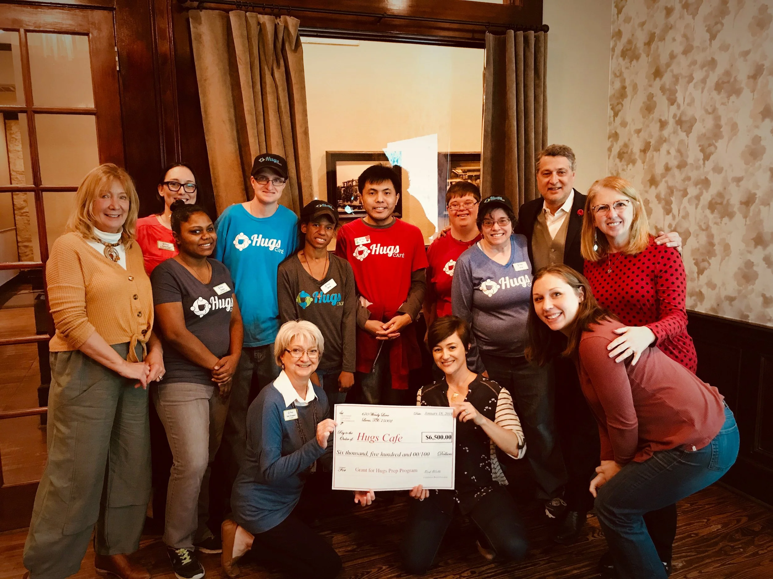 Group of people posing indoors for a photo, holding a large check for $6,500 made out to Hugs Cafe, celebrating a donation or achievement.