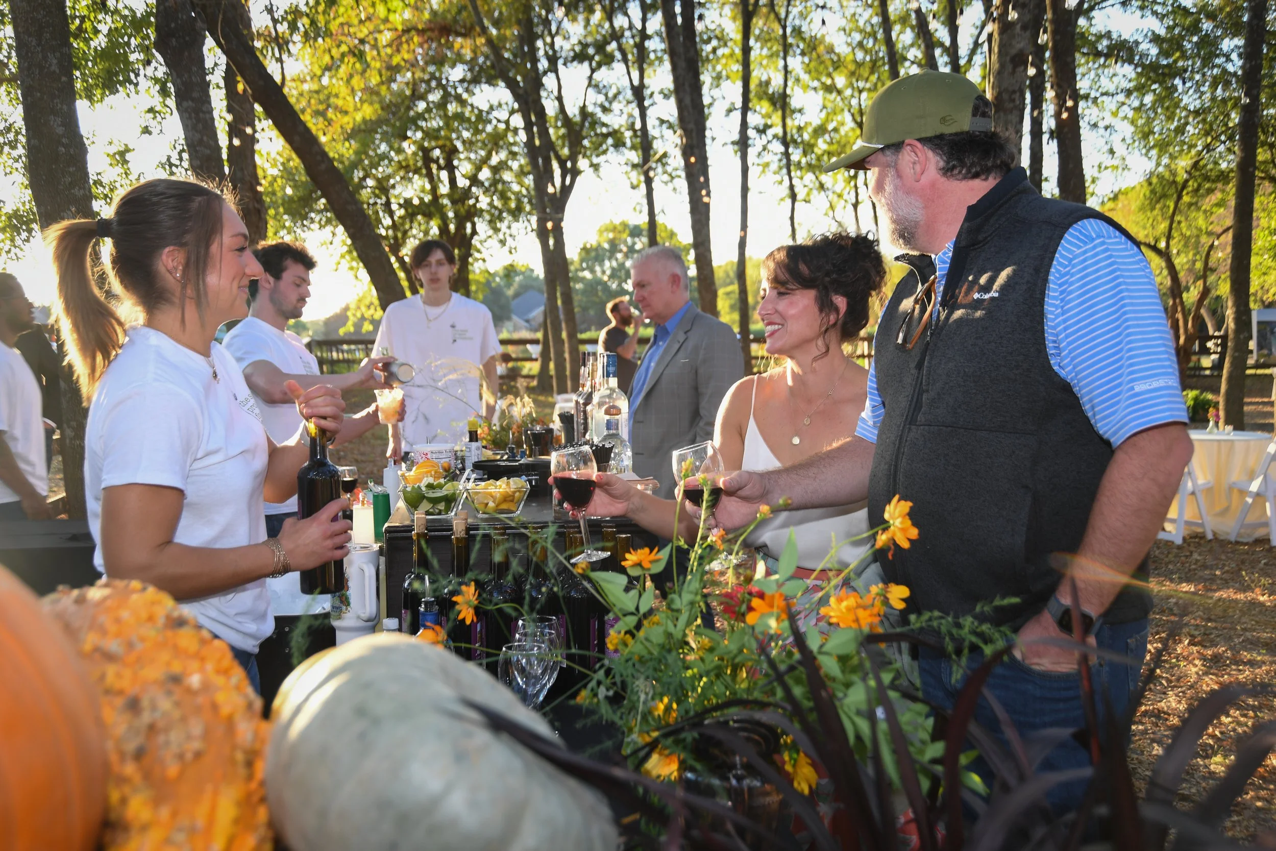 People at an outdoor fall celebration, serving drinks and enjoying each other’s company, with trees and sunlight in the background.