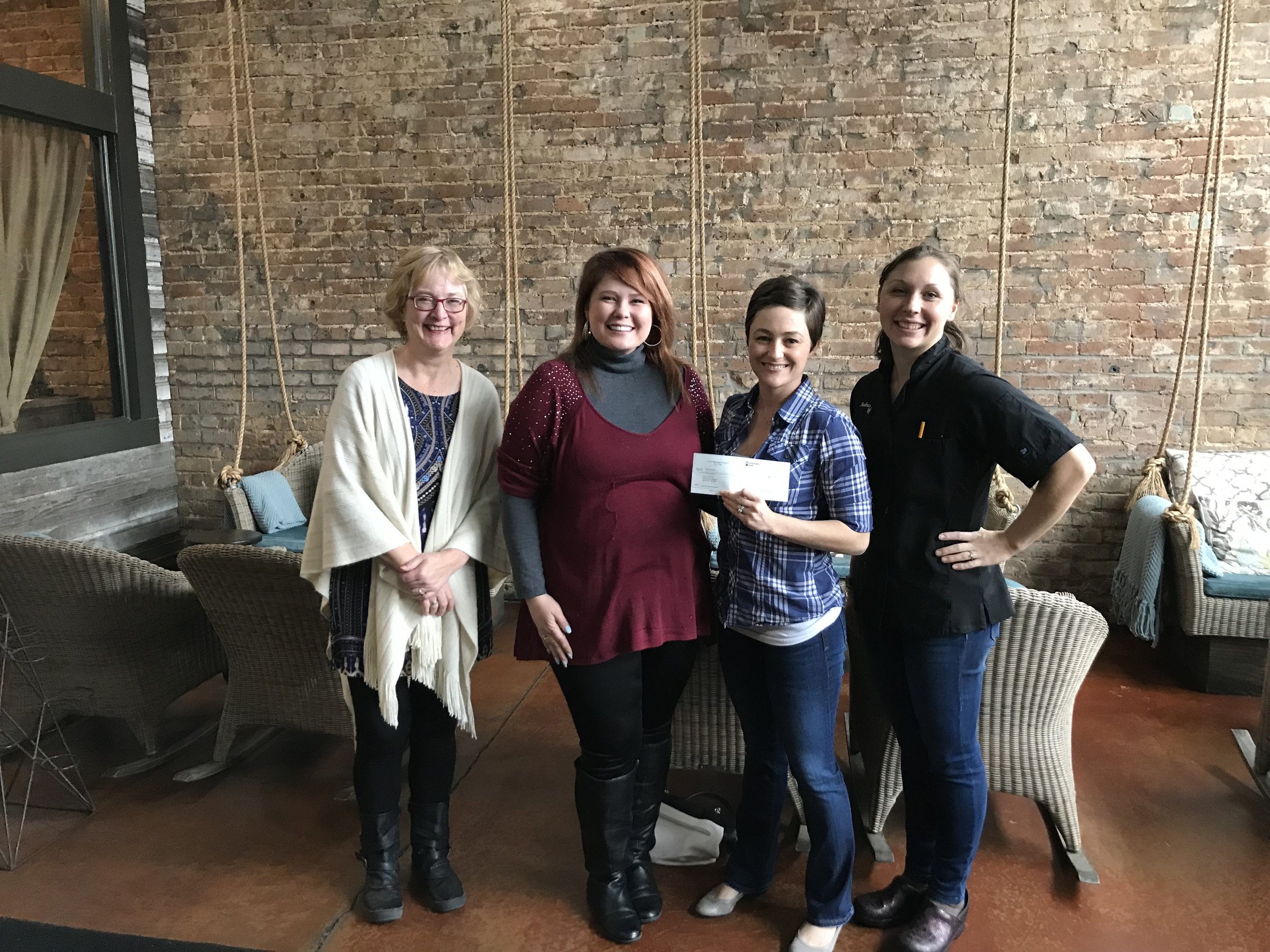 Four women standing together inside a room with a brick wall background, smiling, with the woman second from the right holding a check.
