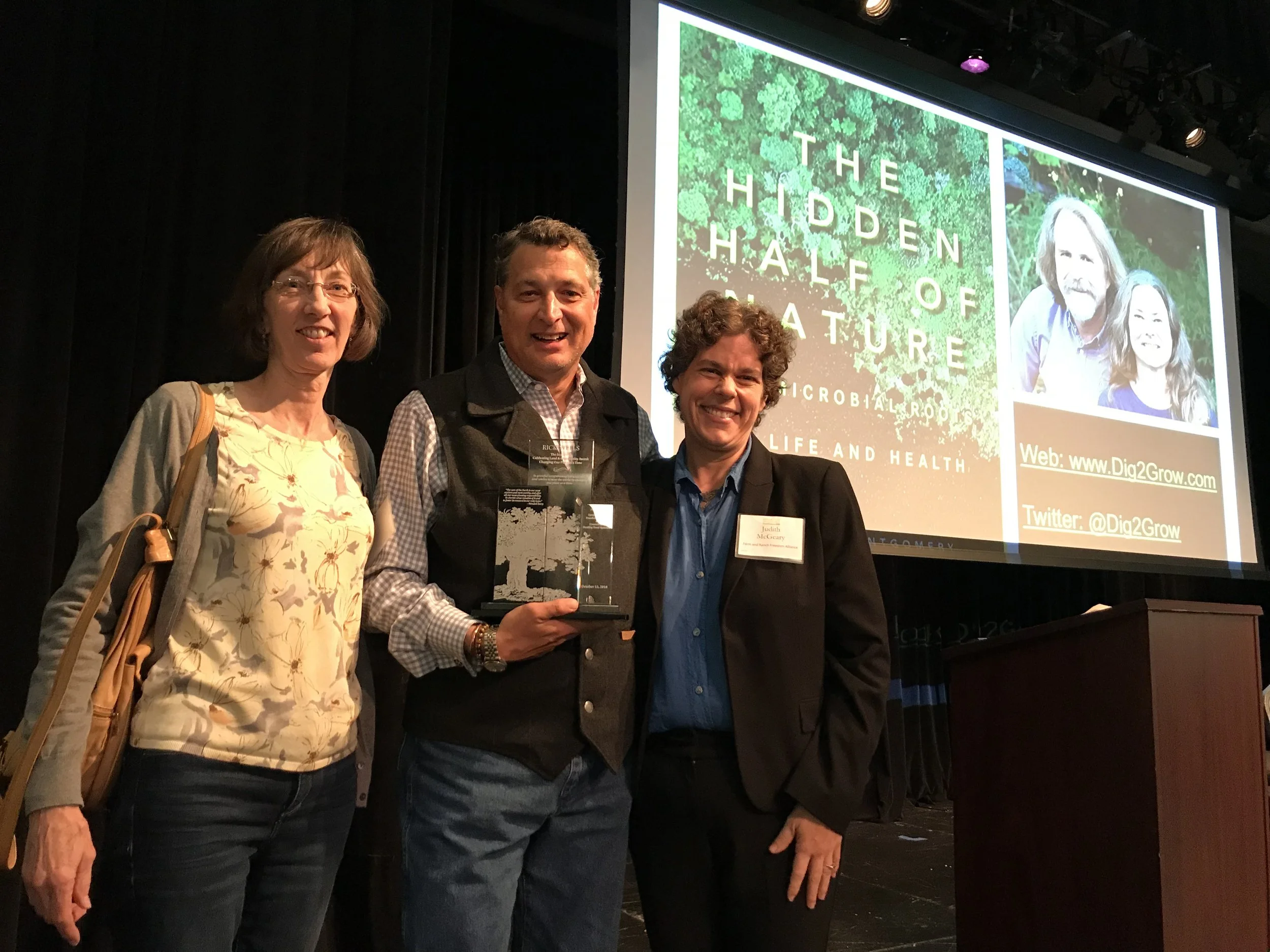Three women and one man standing on stage at an award ceremony. The man, in the center, holds an award. Behind them is a large screen displaying a book cover titled 'The Hidden Half of Nature' and information including a website and Twitter handle.