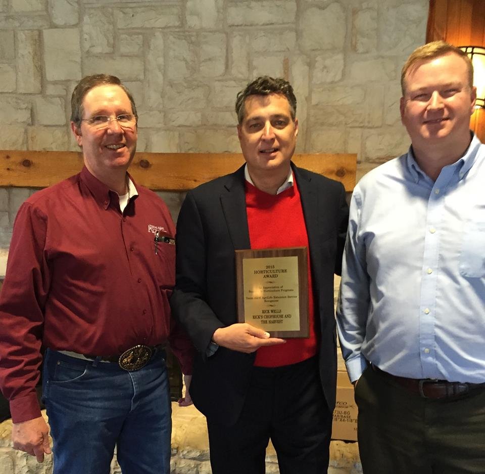 Three men standing together indoors, with the man in the middle holding a plaque award. The background features a stone wall and a wooden beam.
