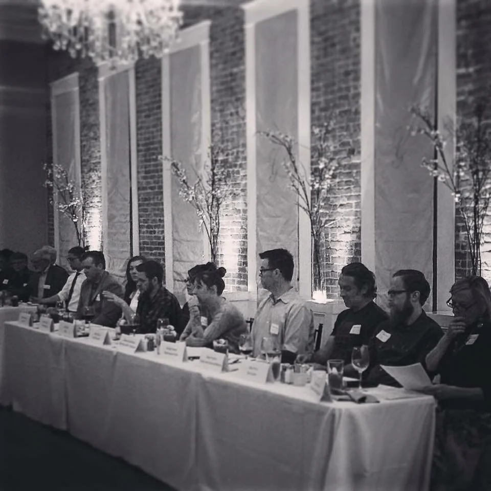 A group of people sitting at a long conference table with name tags, glasses, and papers in a room with exposed brick walls and decorative lighting.