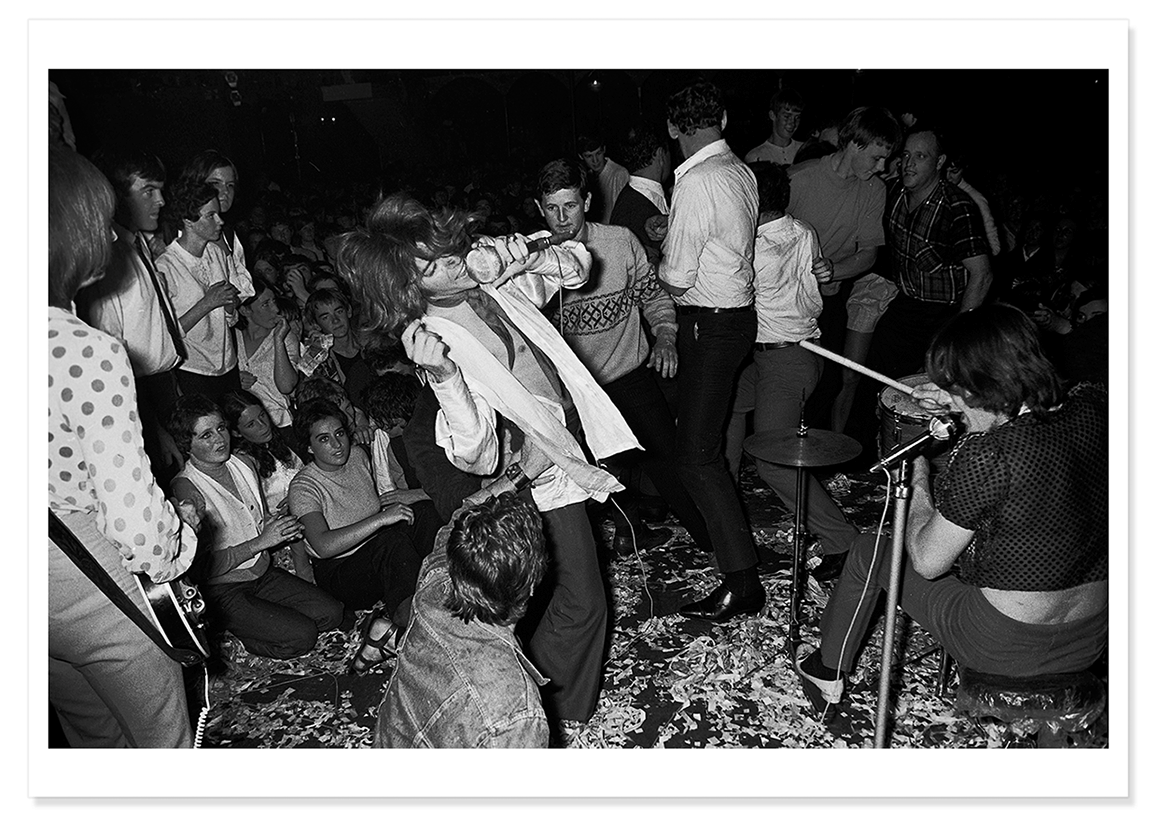 A black and white photograph of a shirtless Jim Keays with light jacket and cravat performing on stage in 1968.  Print shows borders and drop shadow.