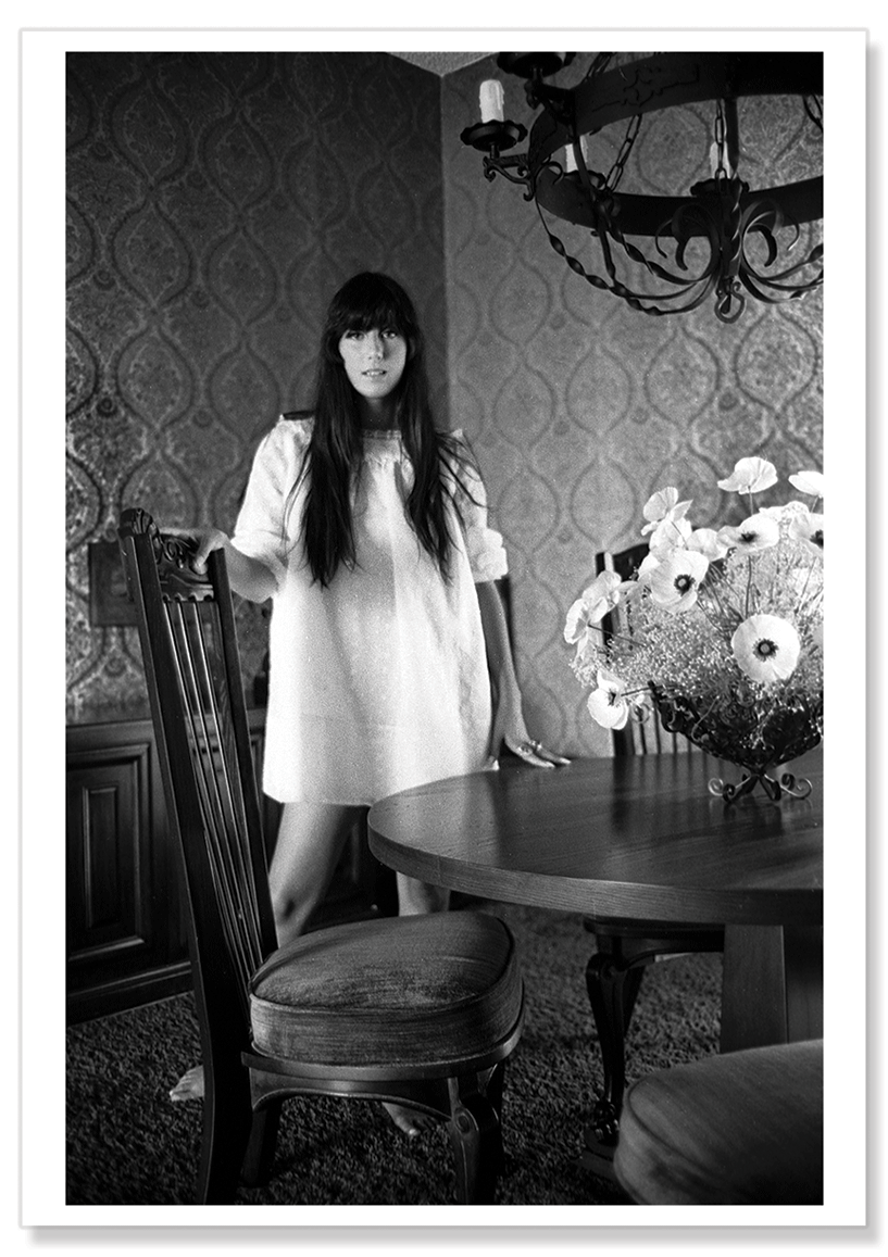 Full-length black and white portrait of Cher in her dining room in Los Angeles in 1967.  Cher is wearing a white smock dress.  Print shows borders and drop shadow.