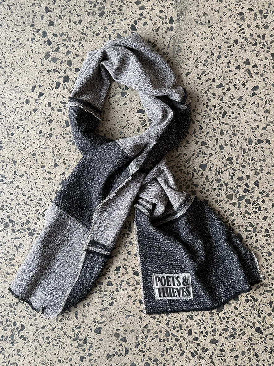 A styled twist of the black and charcoal wool blend scarf with reversed patches and frayed edges, photographed on a concrete floor.