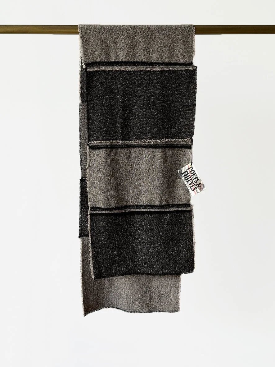 An image of the back of a long slim wool blend scarf with reversed patches and frayed edging in black and charcoal.