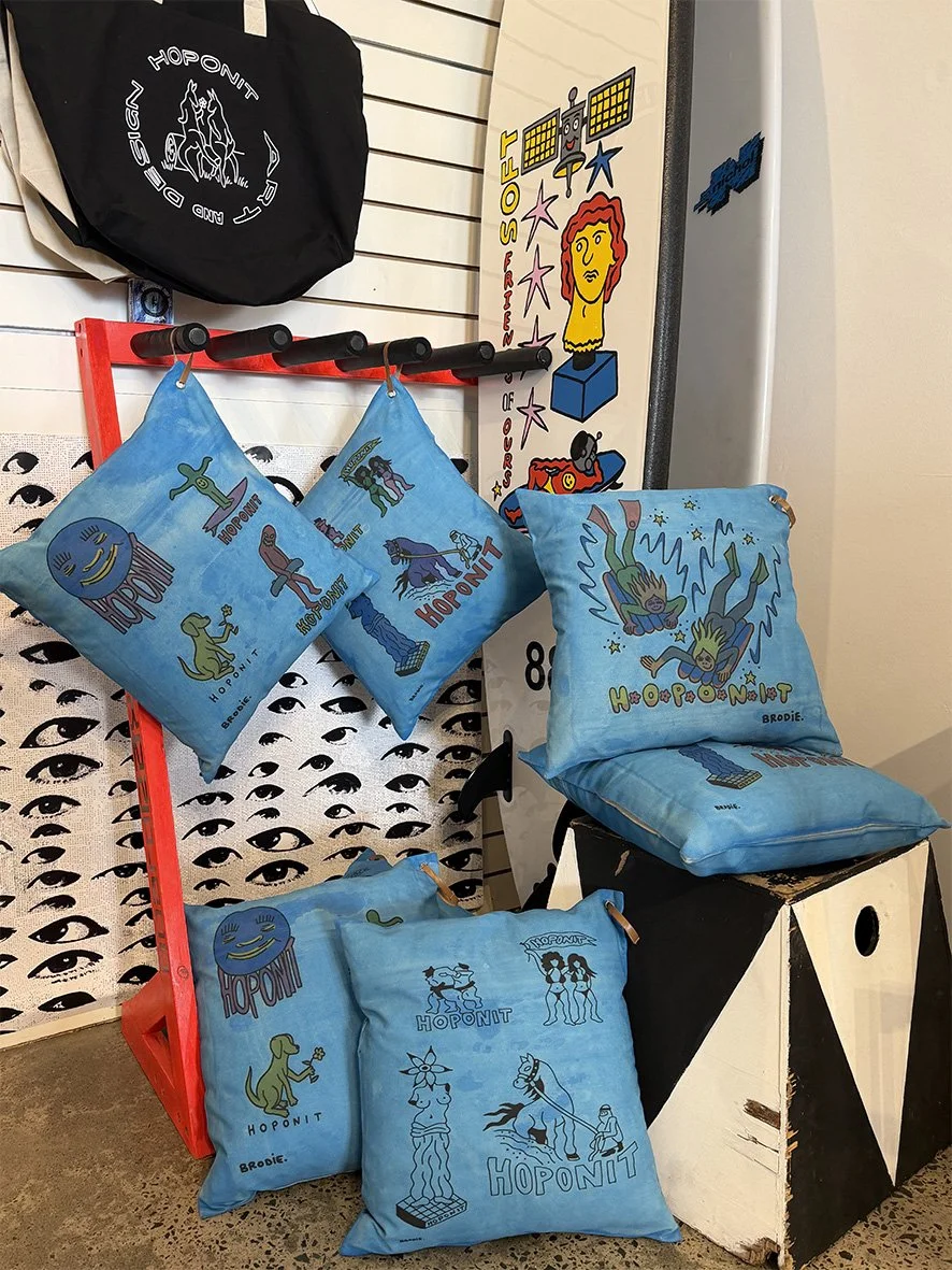 A collection of cushions with sky blue artistic background and images by Brodie Jackson for Hoponit Studio with a skate and surf theme.  A softboard with Brodie Jackson art is in the background.