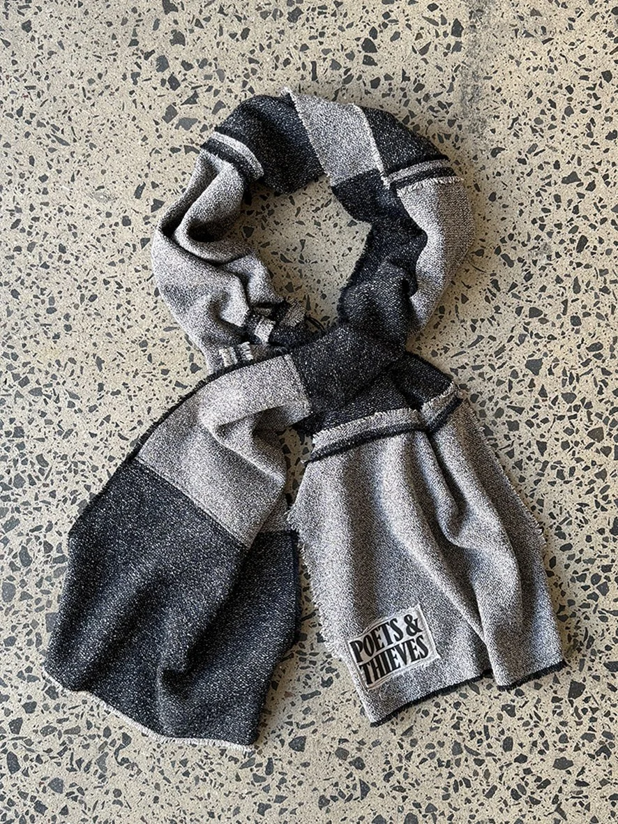 A styled twist of the black and charcoal wool blend scarf with reversed patches and frayed edges, photographed on a concrete floor.