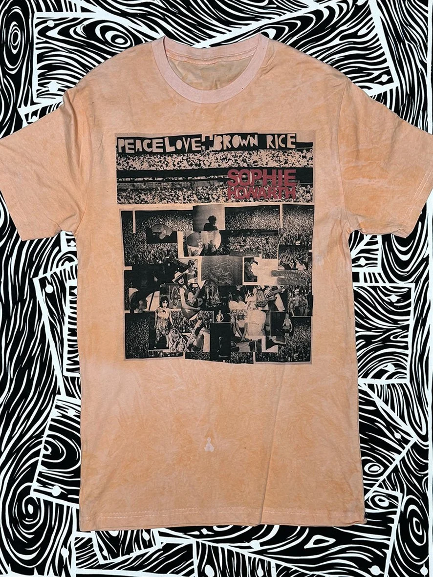 Front view of soft orange Peace Love and Brown Rice tee with a large collage and with black and white woodgrain fabric behind.