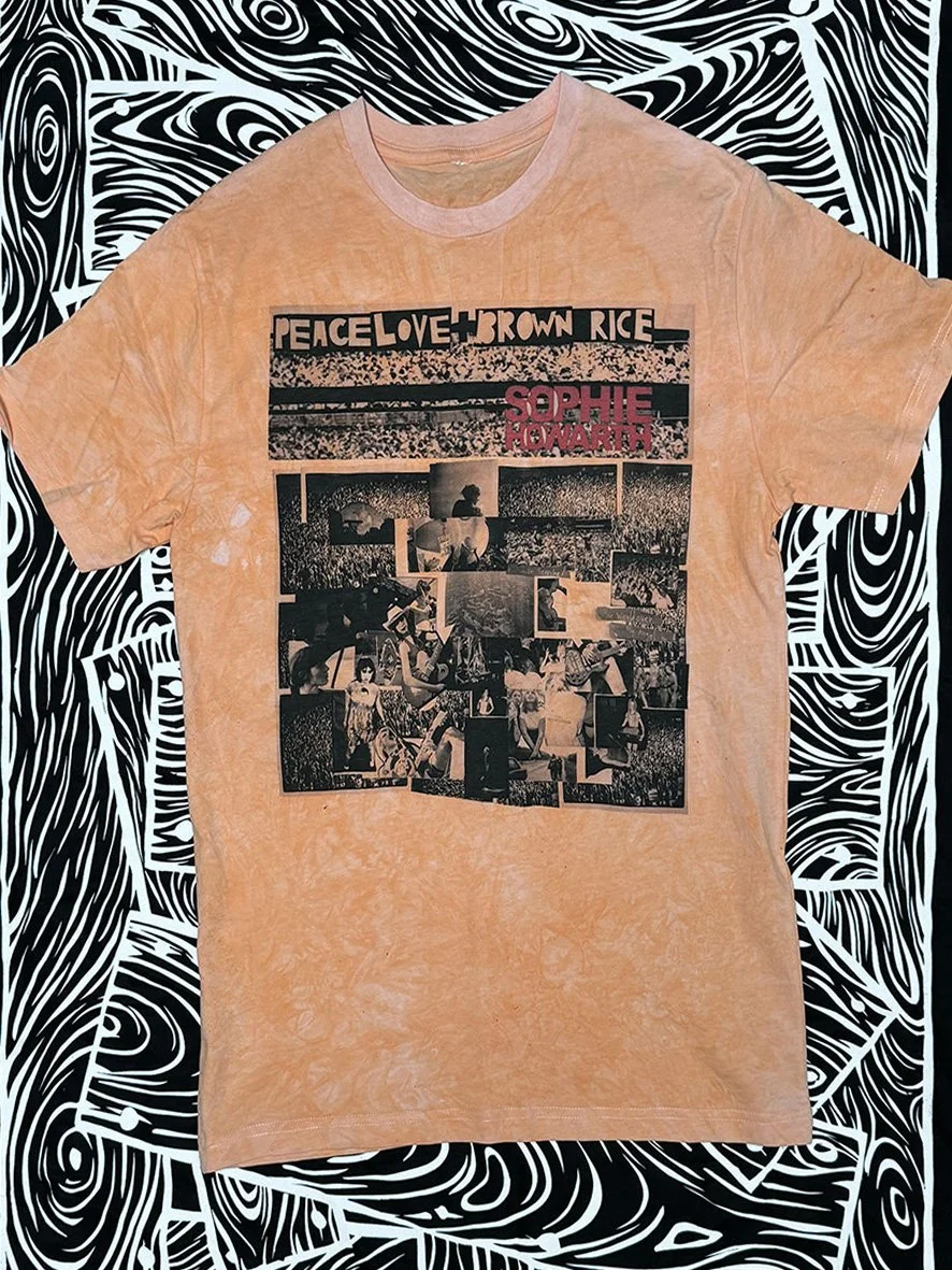 Front view of soft orange Peace Love and Brown Rice tee with a large collage and with black and white woodgrain fabric behind.