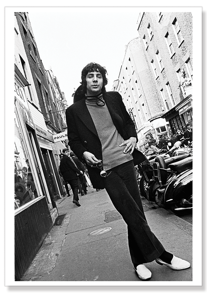 Black and white full length portrait of Cat Stevens in Carnaby Street wearing a turtleneck jumper, velvet pants and white shoes. Print shows borders and drop shadow.