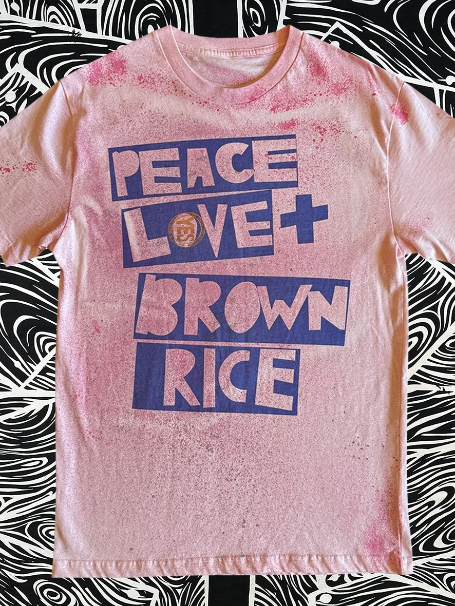 Front view of oxidised pink tee with a blue Peace Love and Brown Rice print and with black and white woodgrain fabric behind.