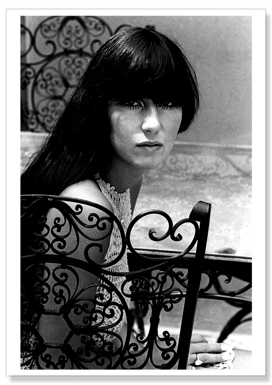 A black and white photographic portrait of Cher sitting outside her home on a chair, looking directly at the camera through her long fringe and dark hair.  Print shows borders and drop shadow.