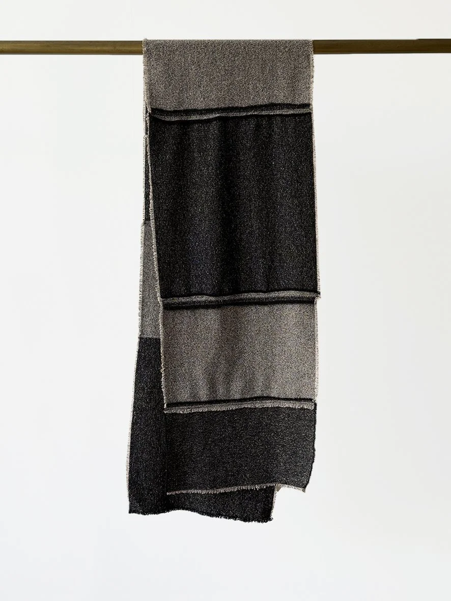 An image of the back of a long slim wool blend scarf with reversed patches and frayed edging in black and charcoal.