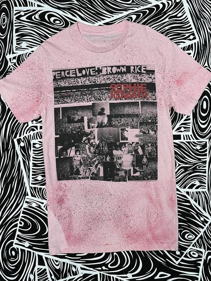 Front view of pink Peace Love and Brown Rice treated tshirt with large Peace Love and Brown Rice print and black and white woodgrain fabric in the background.