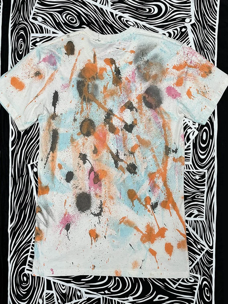 Back view of a natural treated tshirt with orange, pink and soft blue ink and with black and white woodgrain fabric in the background.