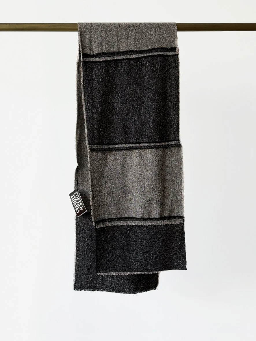 An image of the back of a long slim wool blend scarf with reversed patches and frayed edging in black and charcoal.