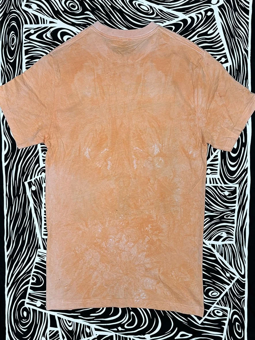 Back view of soft orange Peace Love and Brown Rice tee with black and white woodgrain fabric behind.