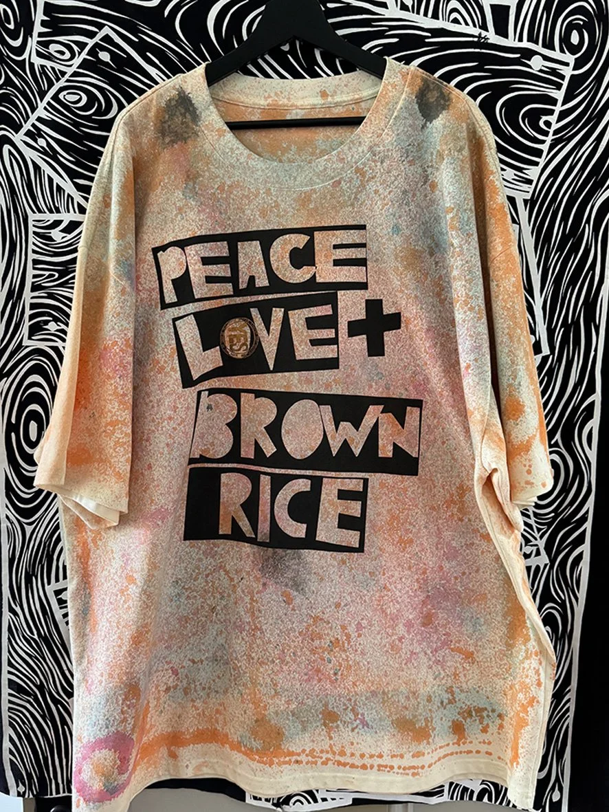 Peace Love and Brown Rice heavy oversized tshirt with oxidised treatment hanging in front of a black and white woodgrain fabric.