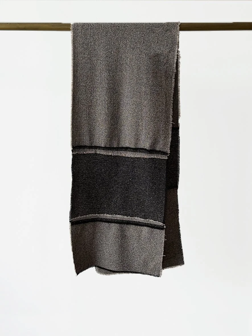 An image of the back of a long slim wool blend scarf with reversed patches and frayed edging in black and charcoal.