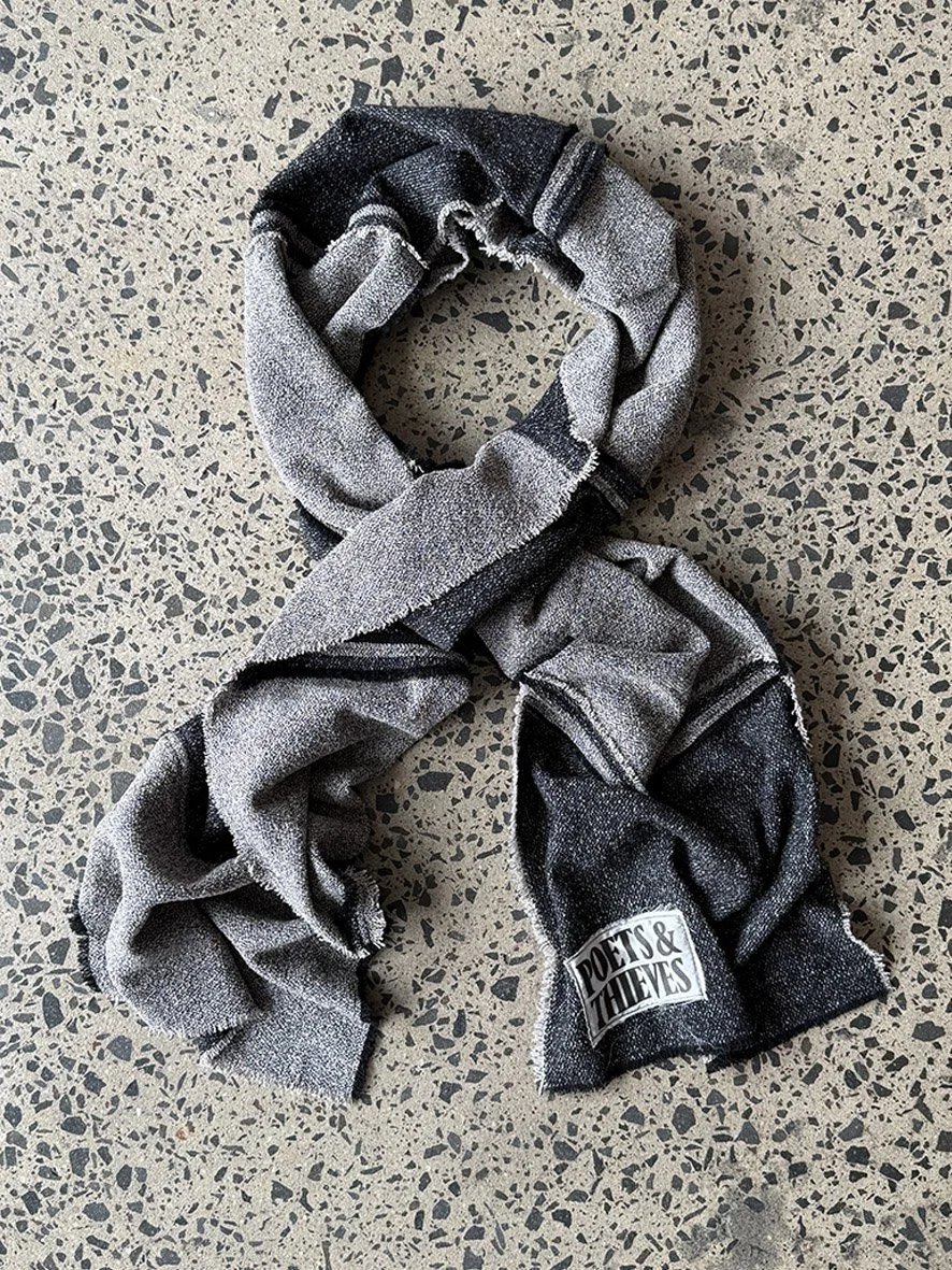 A styled twist of the black and charcoal wool blend scarf with reversed patches and frayed edges, photographed on a concrete floor.