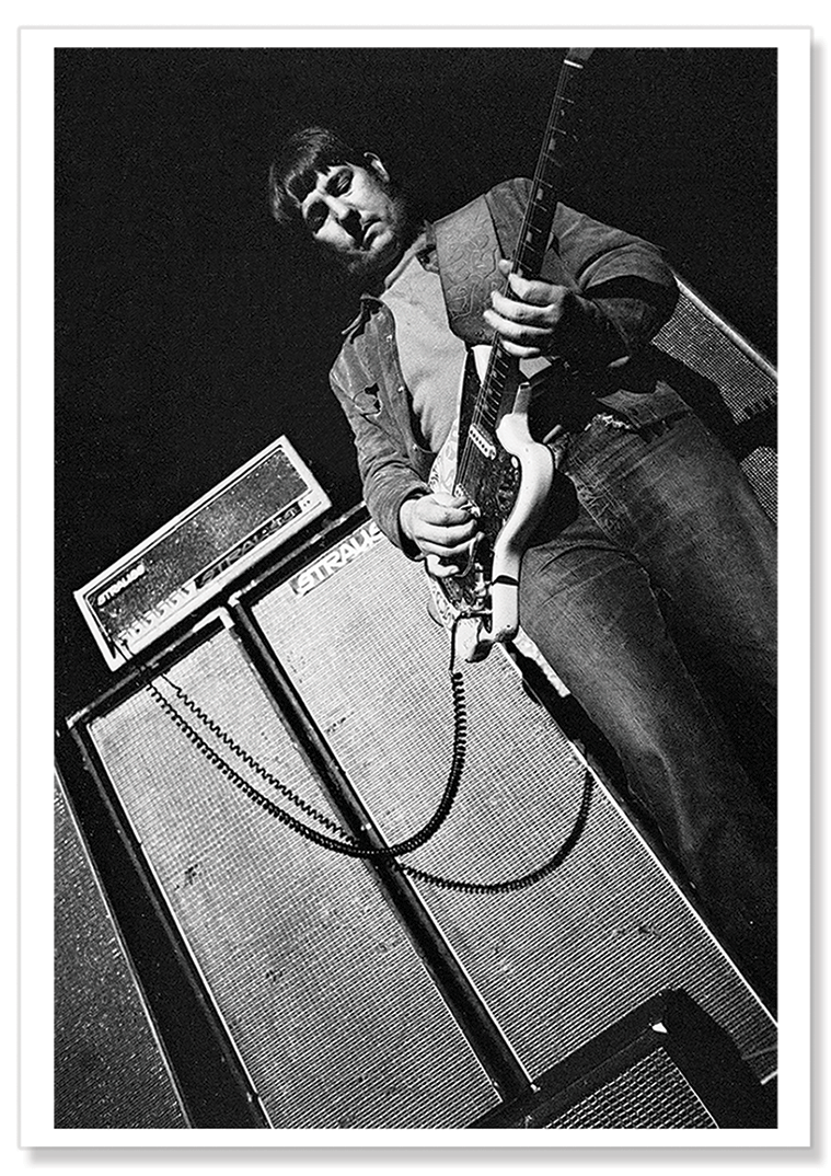 Black and white photographic portrait taken from below of Lobby Loyde playing guitar with acoustic equipment behind.  Print shows borders and drop shadow.