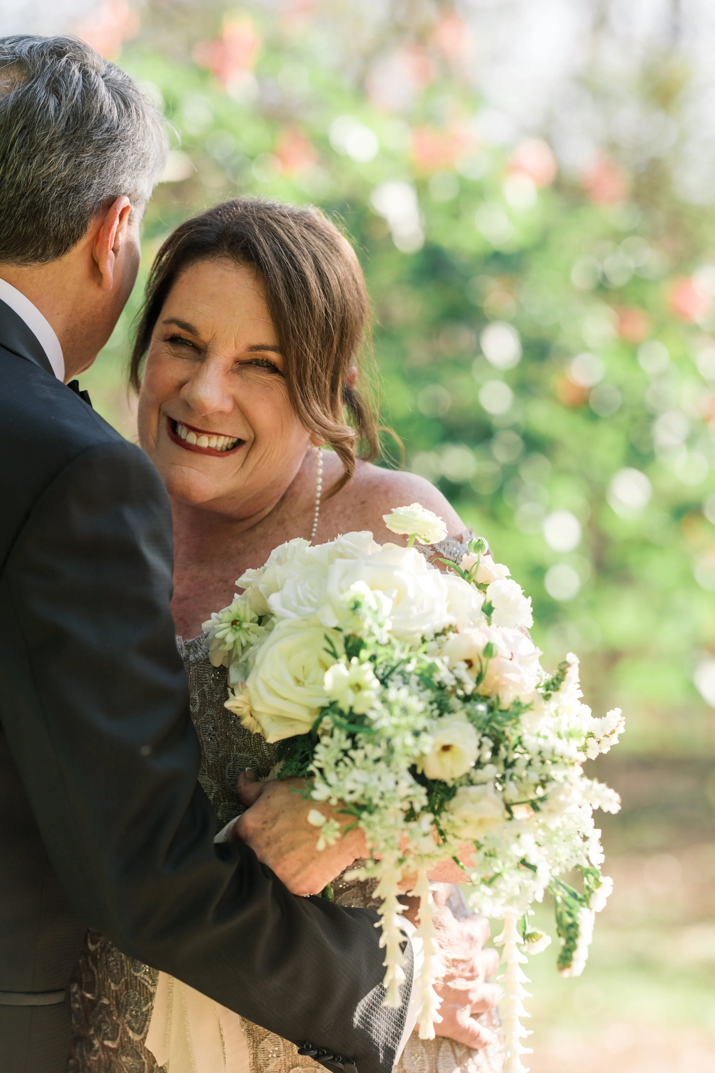 A bride holding a bouquet of white and green flowers, smiling outdoors.