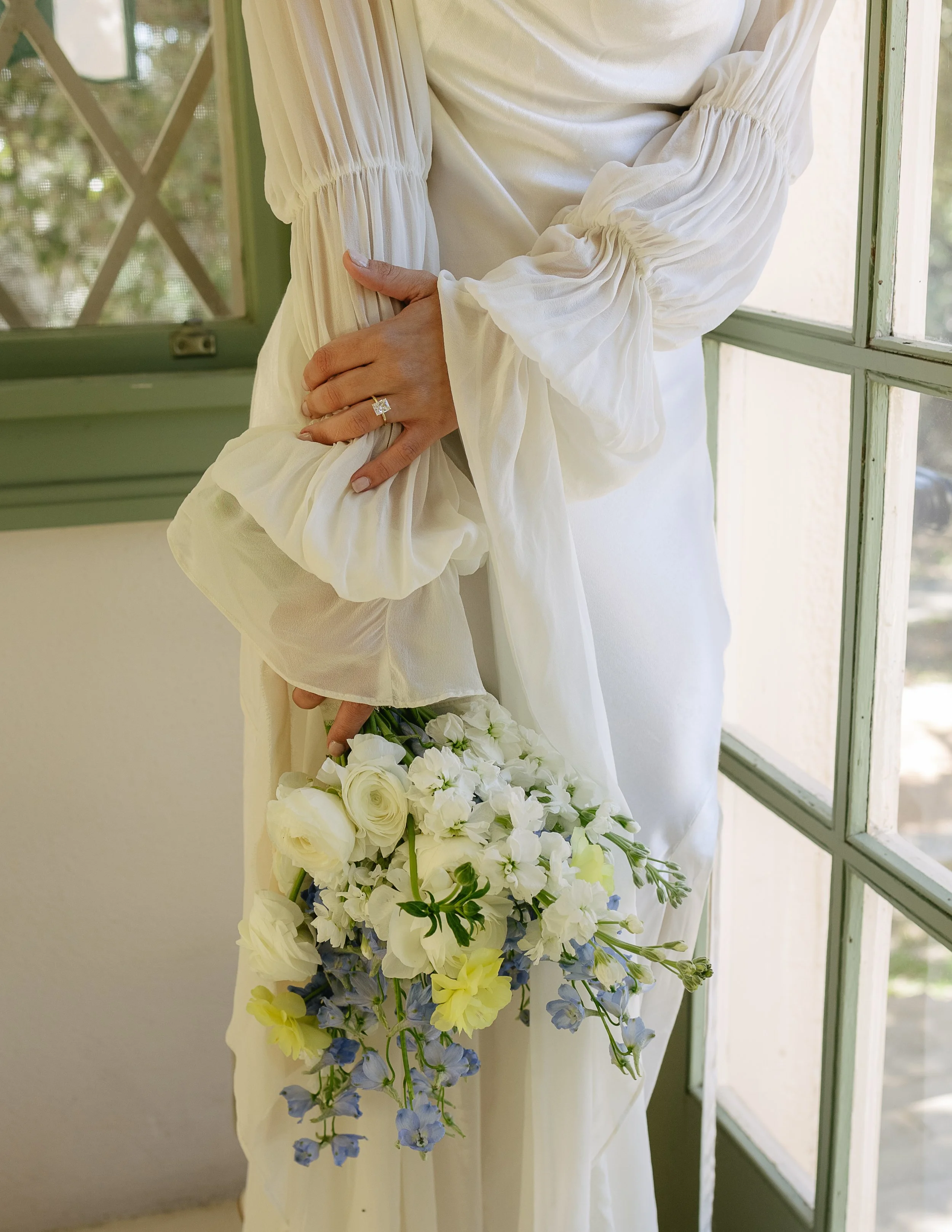 Person in a white dress holding a bouquet of white and blue flowers near a window.