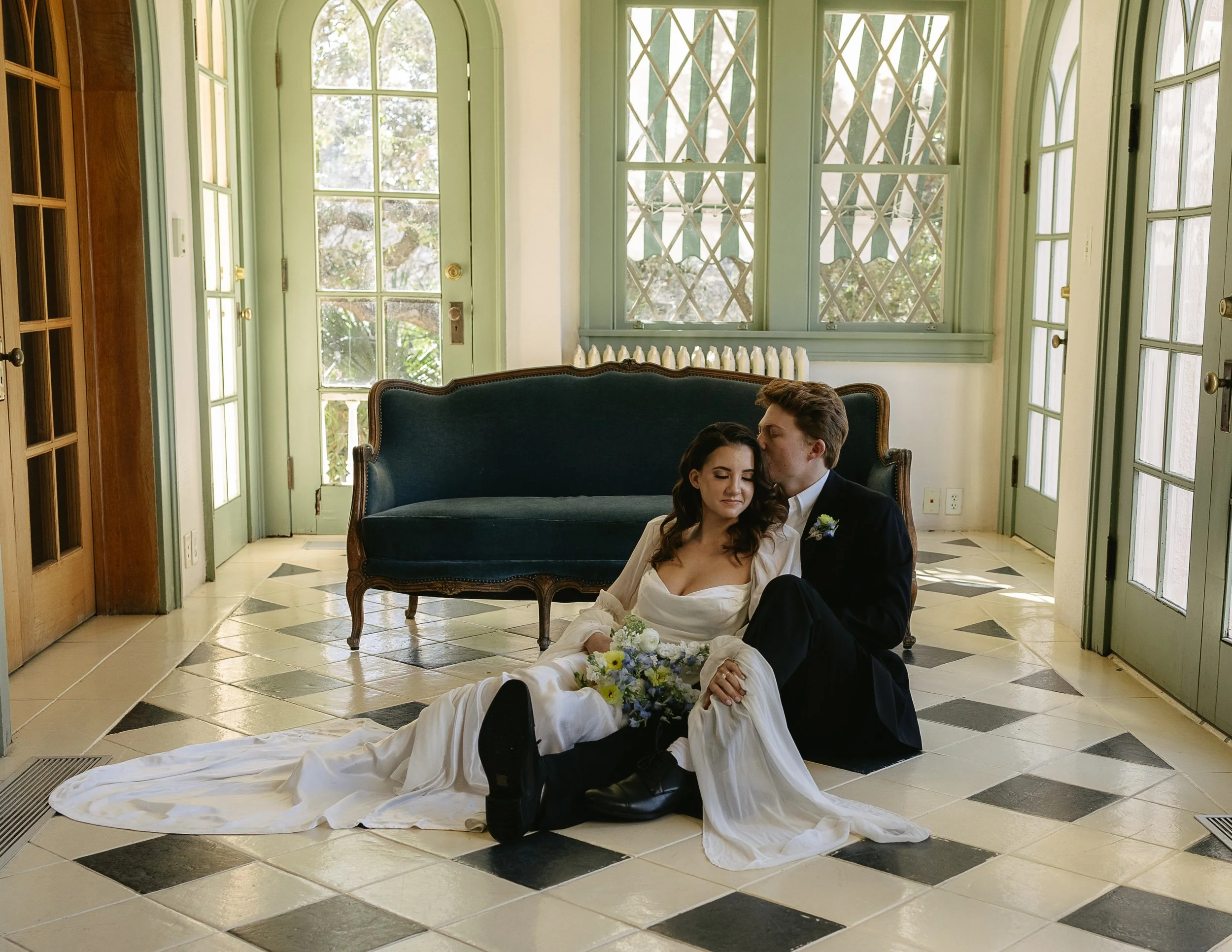 A couple in wedding attire seated on a checkered floor, leaning against a vintage couch in a sunlit room.