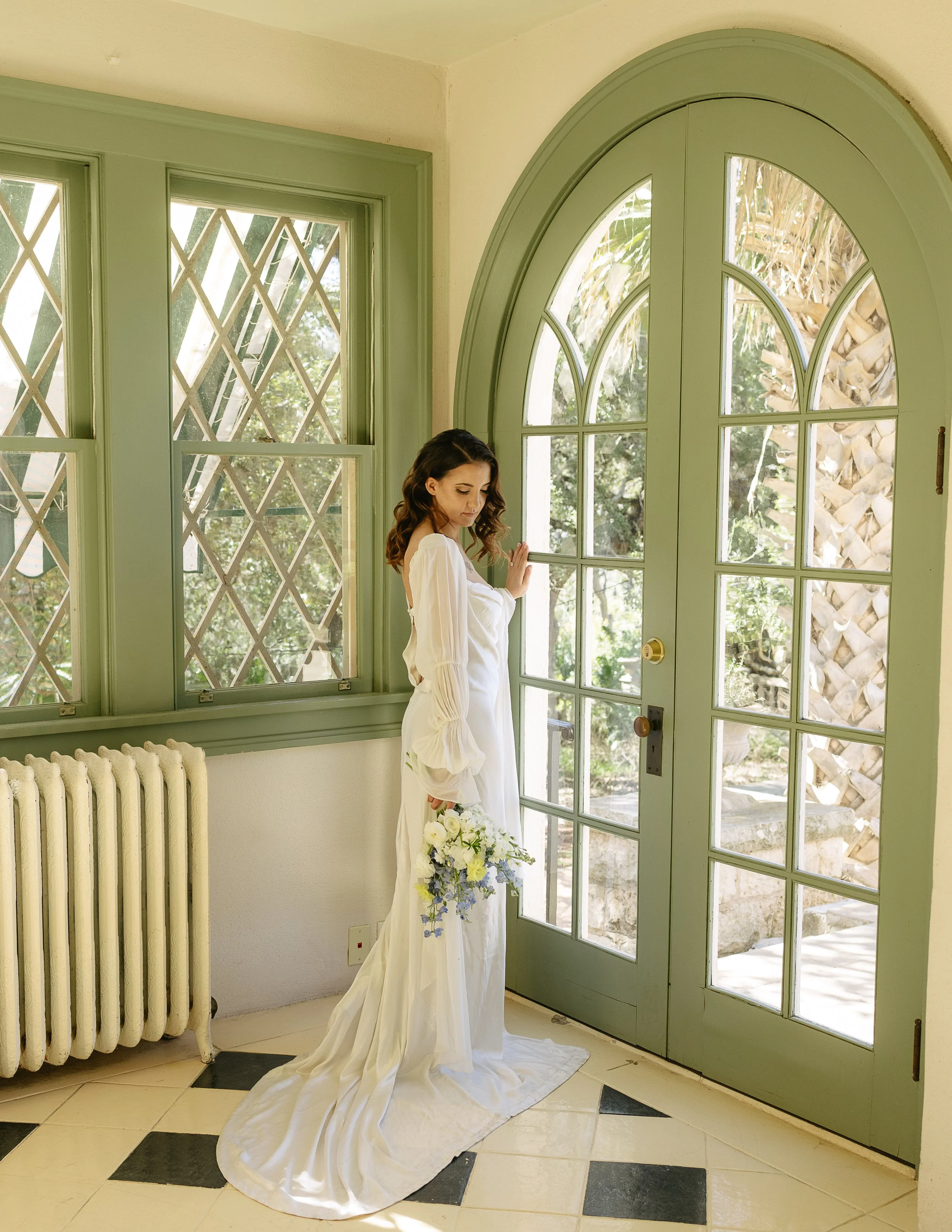Bride in white gown holding bouquet, standing by green French doors and windows.