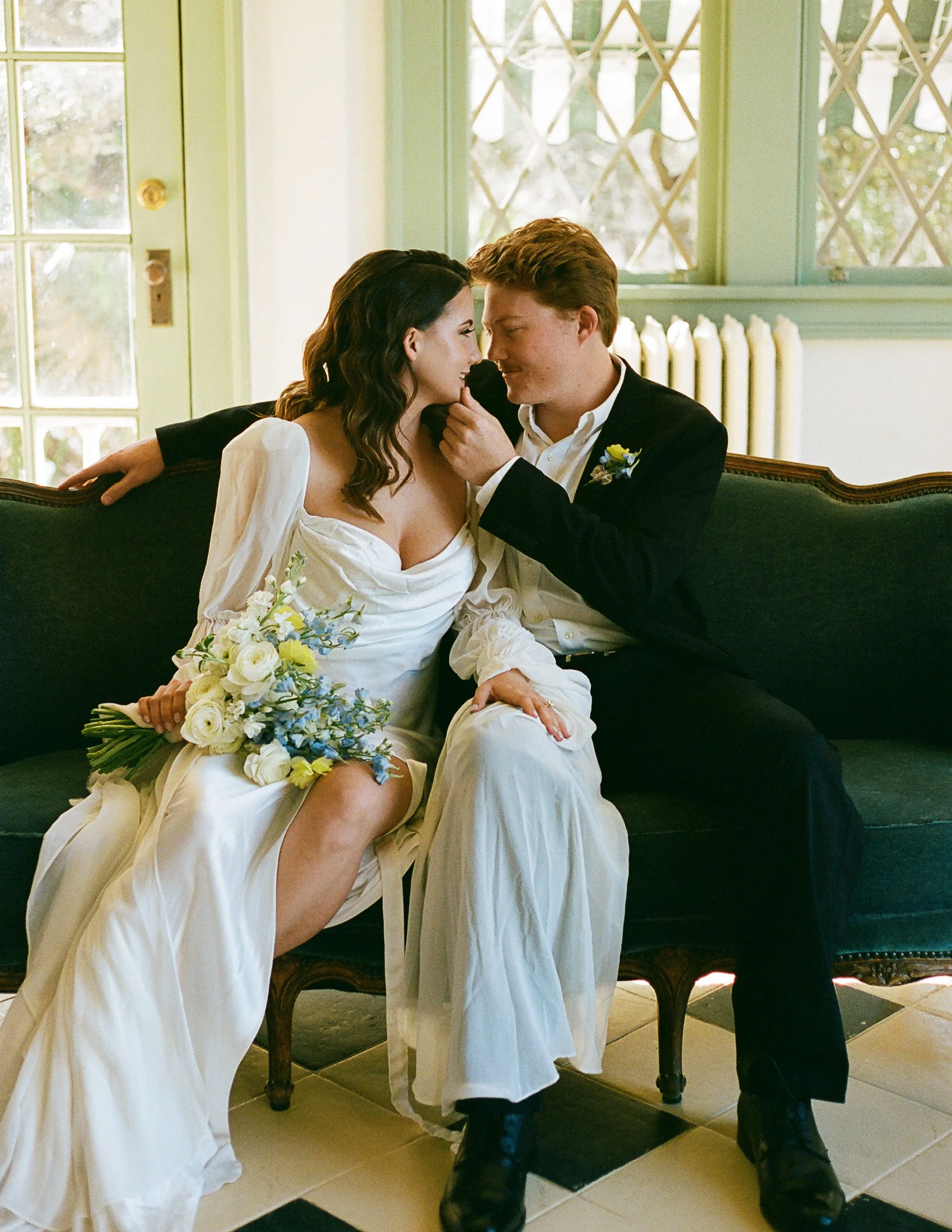 A couple sitting on a couch, embracing. The woman wears a white dress, holding a bouquet of flowers, and the man is in a suit. They are in a room with green-framed windows.