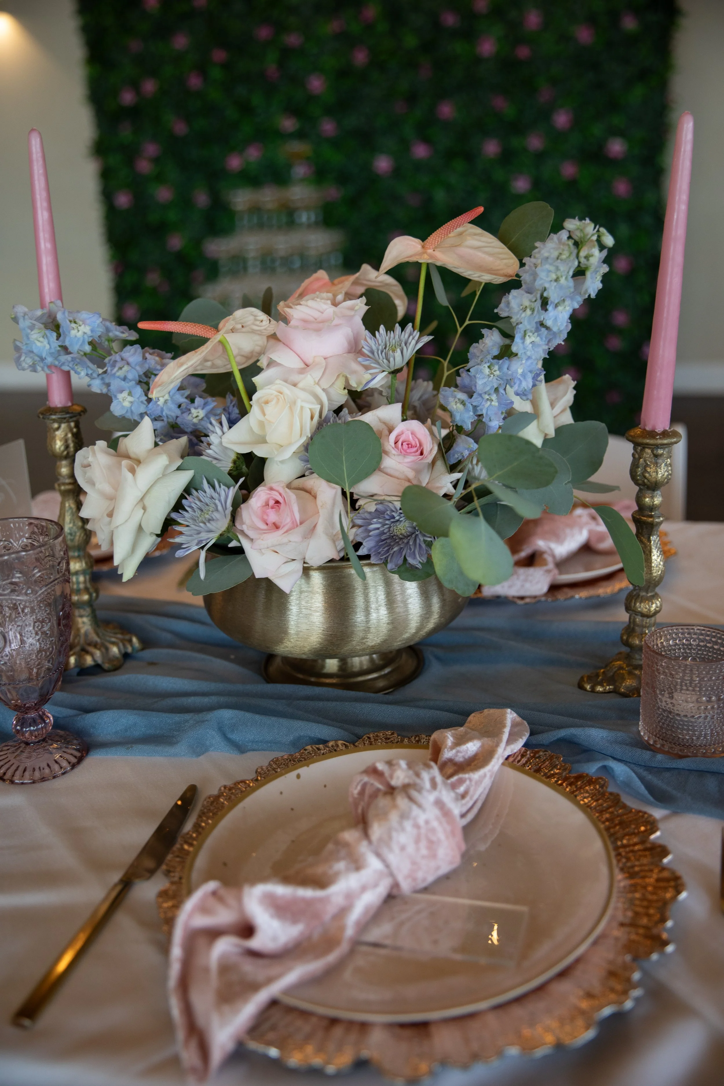 Elegant table setting with gold-rimmed plates, pink and purple floral centerpiece, pink candles, and textured glassware on blue and white linens.