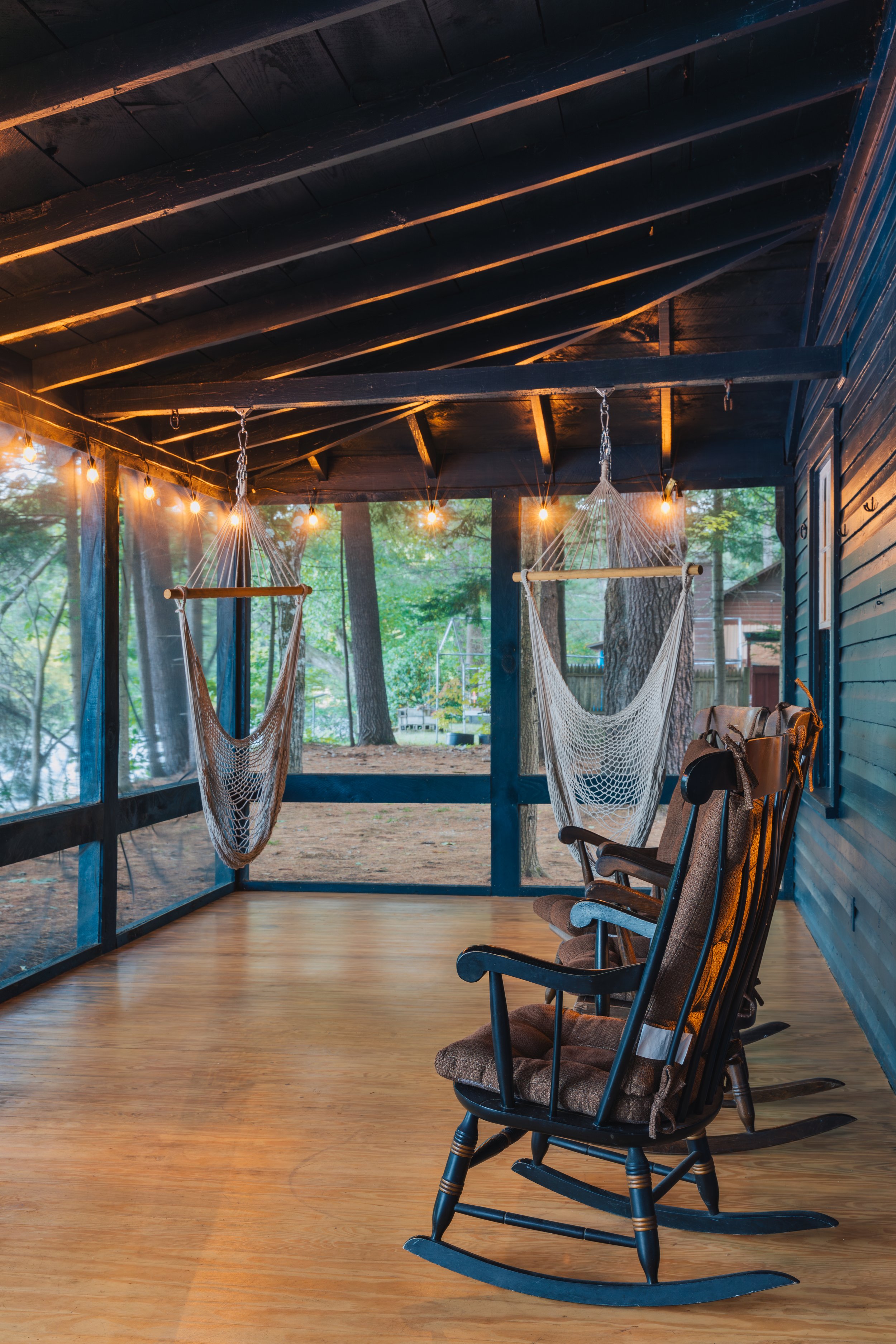 Porch with wooden floor, three rocking chairs, hanging hammock chairs, string lights, surrounded by trees and greenery.