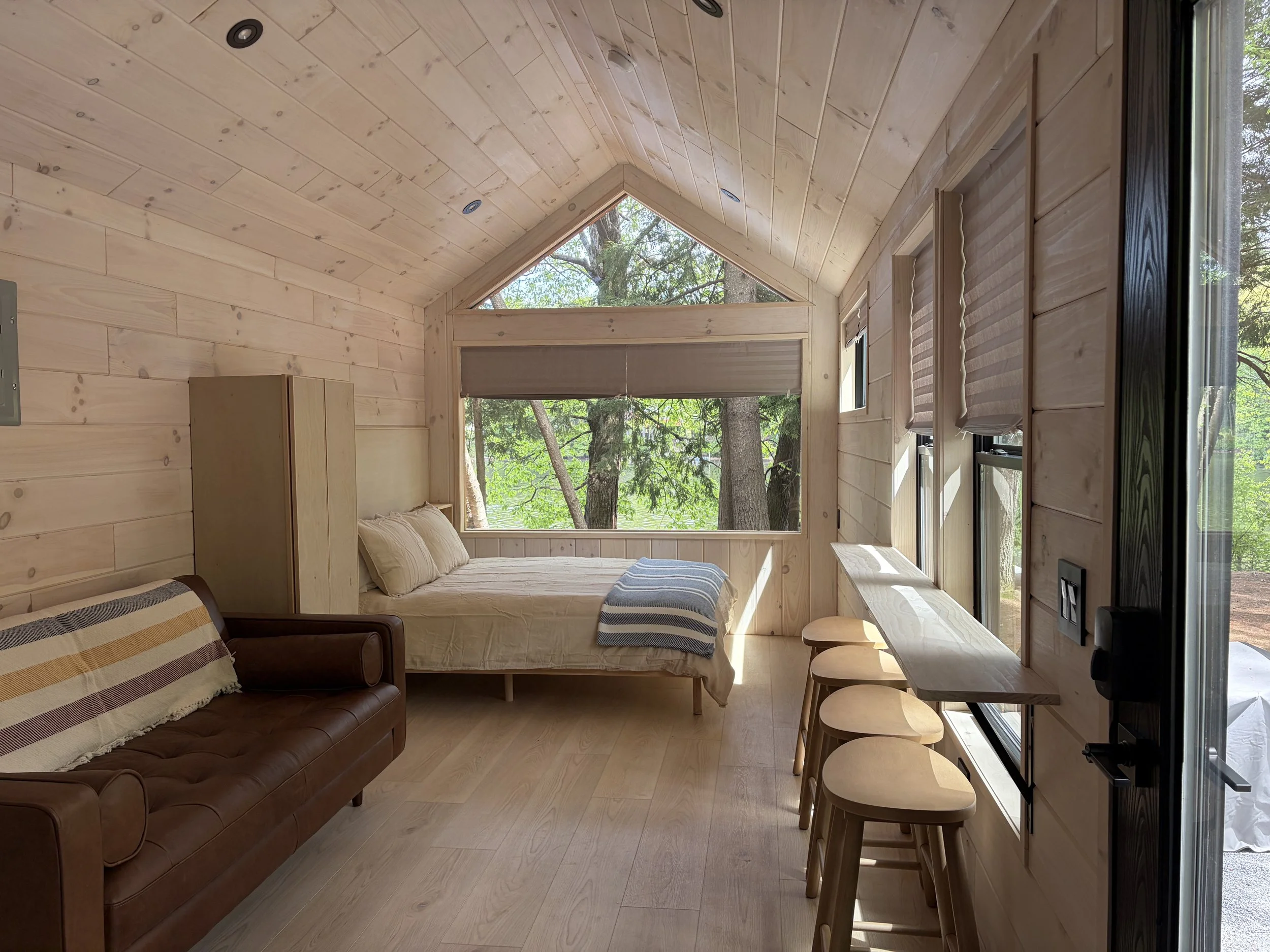 Interior of a cozy cabin with wood panel walls and ceiling, a bed with cream bedding, a brown leather sofa with a striped blanket, and bar stools next to a counter with windows showing trees outside. This is Adirondack living at its best.