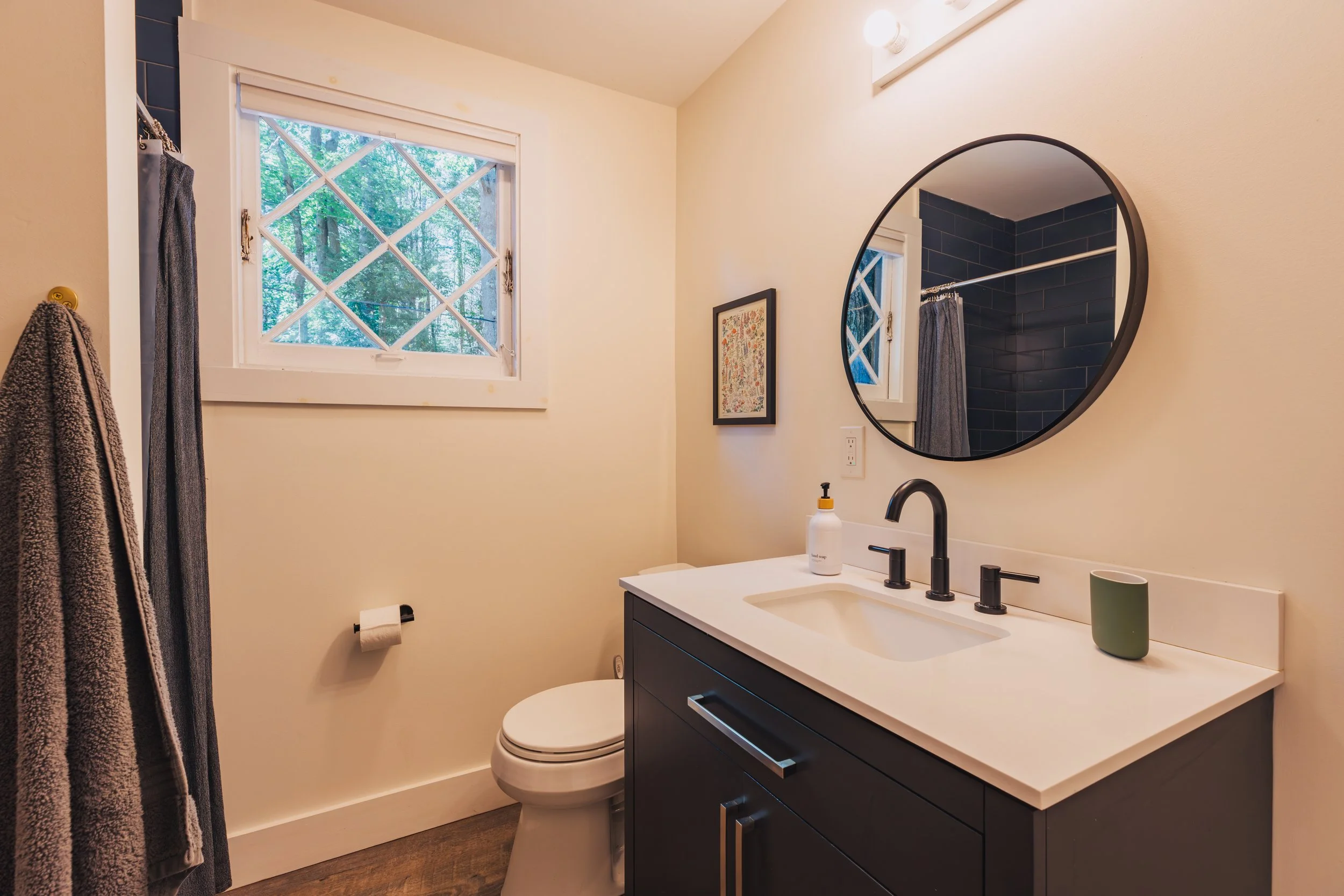 A bathroom with a white wall, a window with a view of trees, a round mirror above a dark vanity with a white countertop, a black faucet, a soap dispenser, a green cup, a towel hanging on a hook, and a toilet.