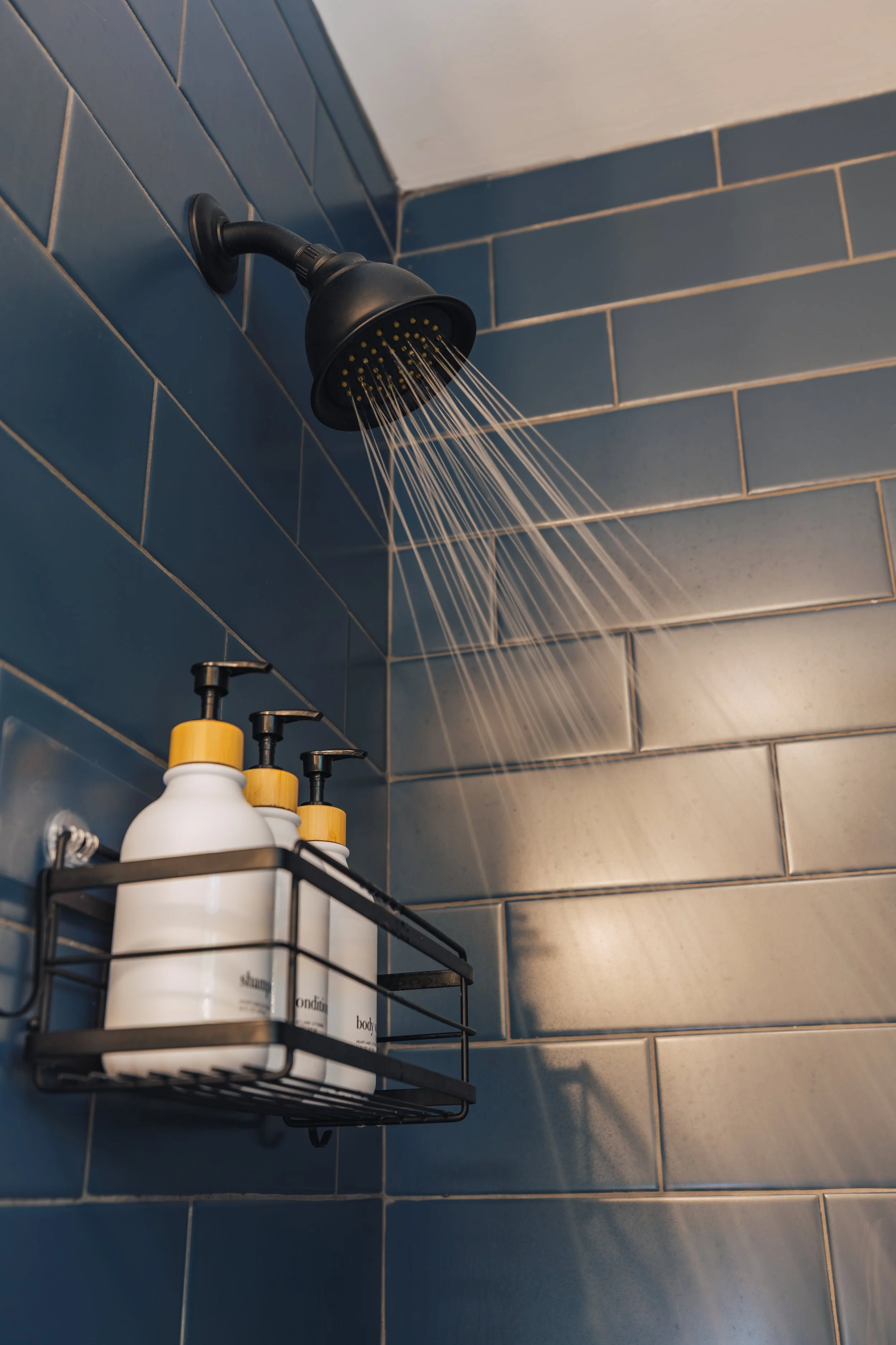 A black showerhead mounted on a blue tiled wall in a shower, with water spraying out. Below it, a metal shelf holds three white bottles with black pumps and yellow accents.