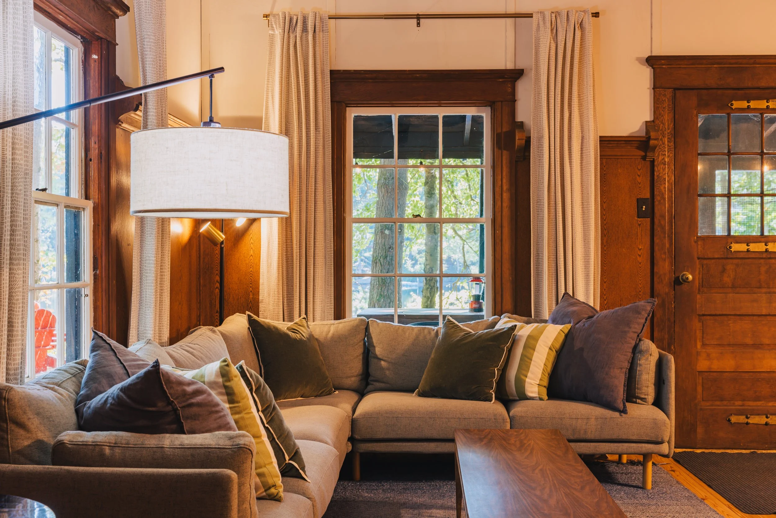 A cozy living room with a beige sectional sofa adorned with various green and striped pillows. Behind the sofa are large windows with wooden frames, with curtains drawn to the sides, revealing a view of trees outside. A wooden coffee table is in fron