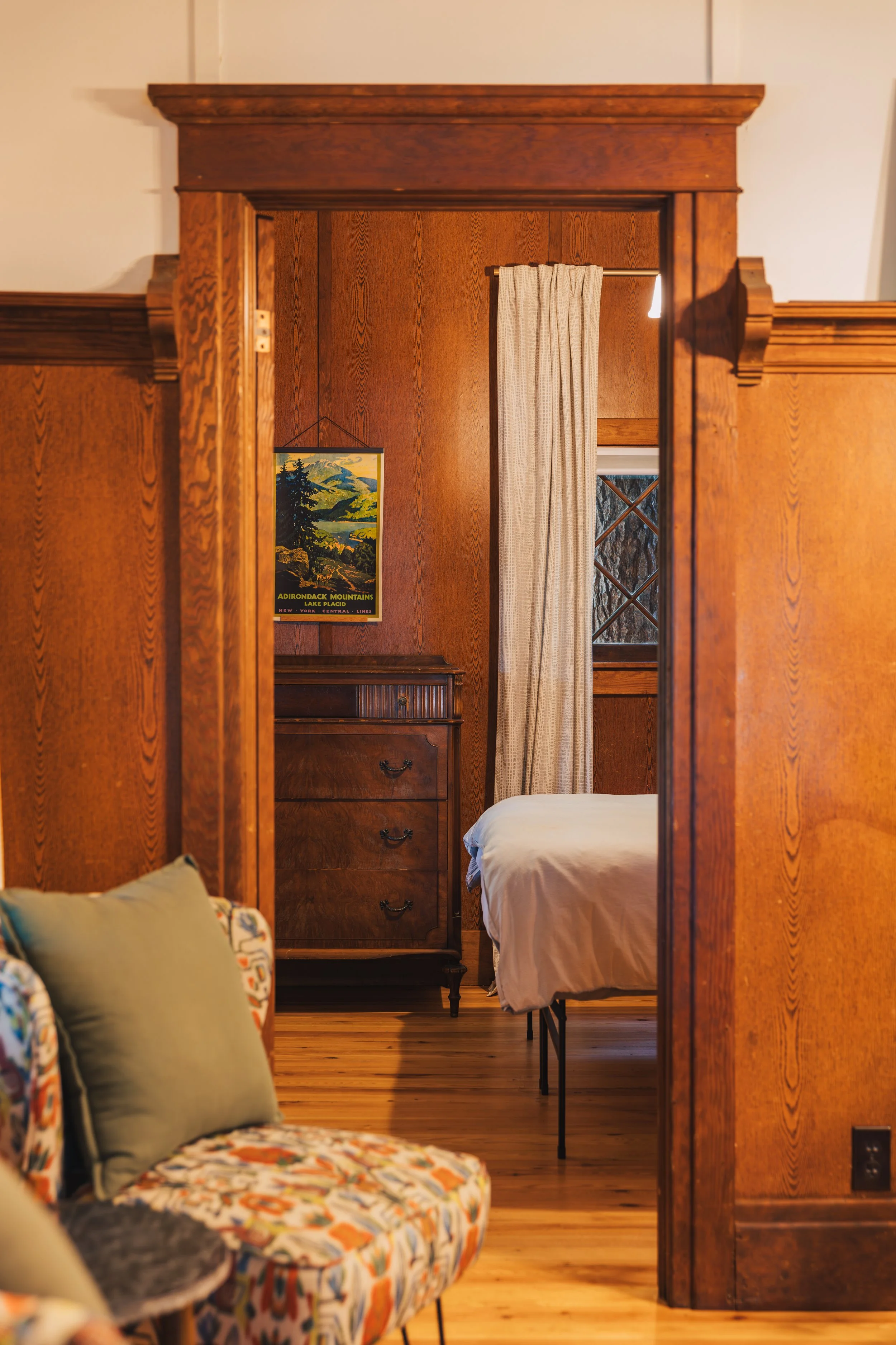 A view through a doorway into a bedroom with wood-paneled walls. The bed is made with white bedding. A window with cream-colored curtains is partially visible, along with a wooden dresser. A vintage travel poster hangs on the wall, and part of a colo