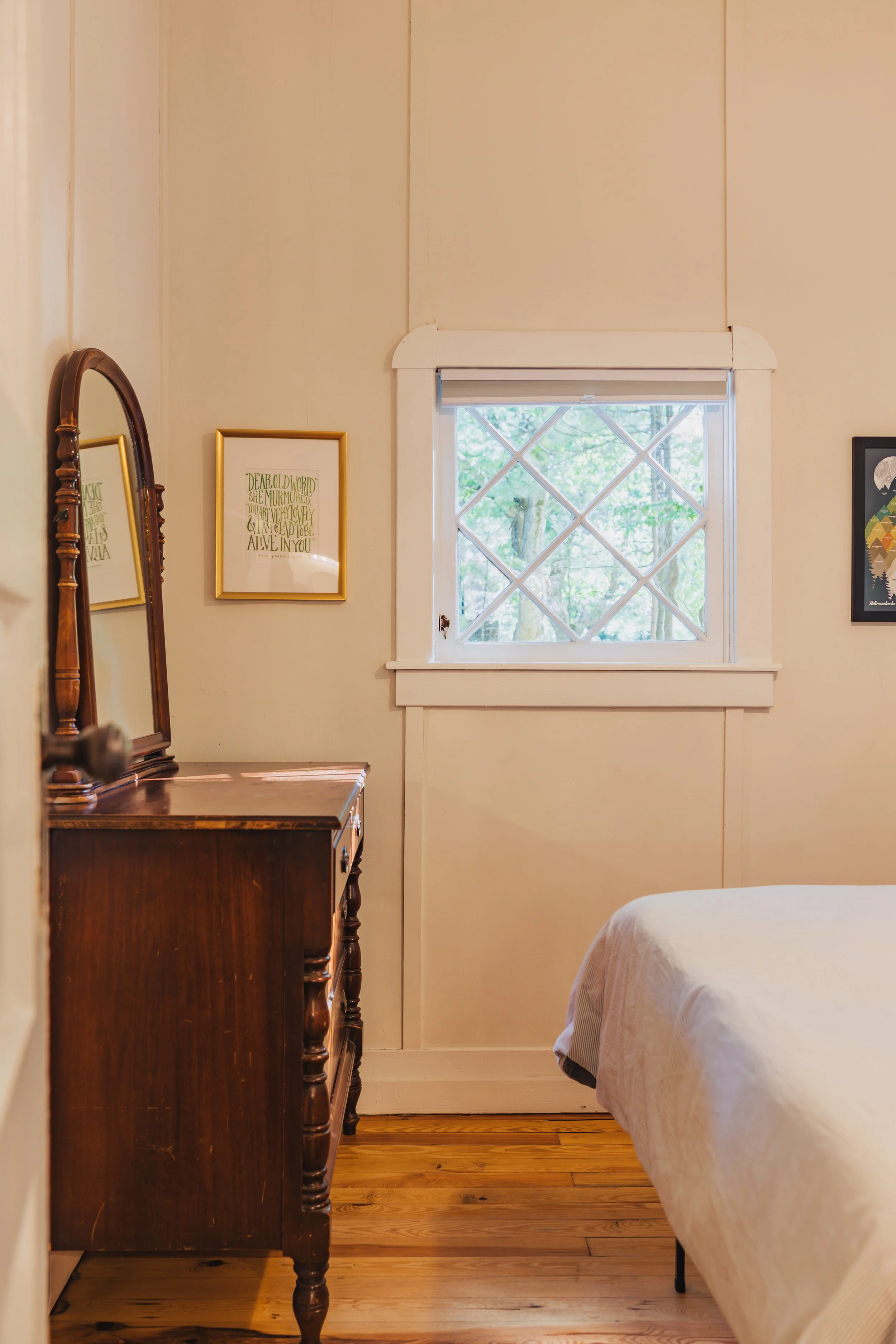 Interior view of a bedroom corner with a wooden dresser, a framed mirror, two picture frames on the wall, a window with a grid design showing trees outside, and part of a bed with white bedding.