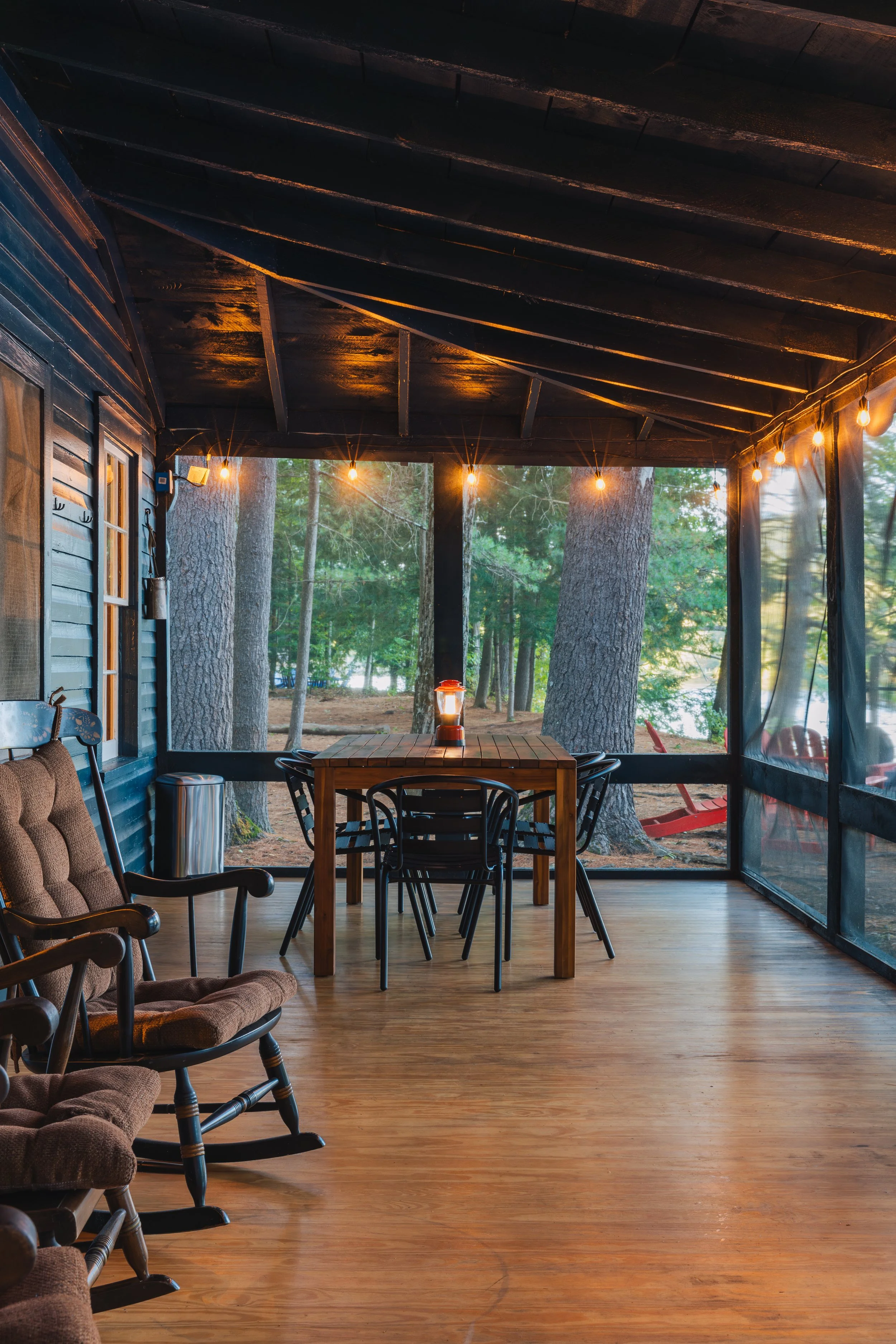 Screen porch with wooden floor and outdoor furniture, including rocking chairs and a rectangular table with chairs, illuminated by string lights, overlooking a wooded area with the Hudson River in the Adirondacks.