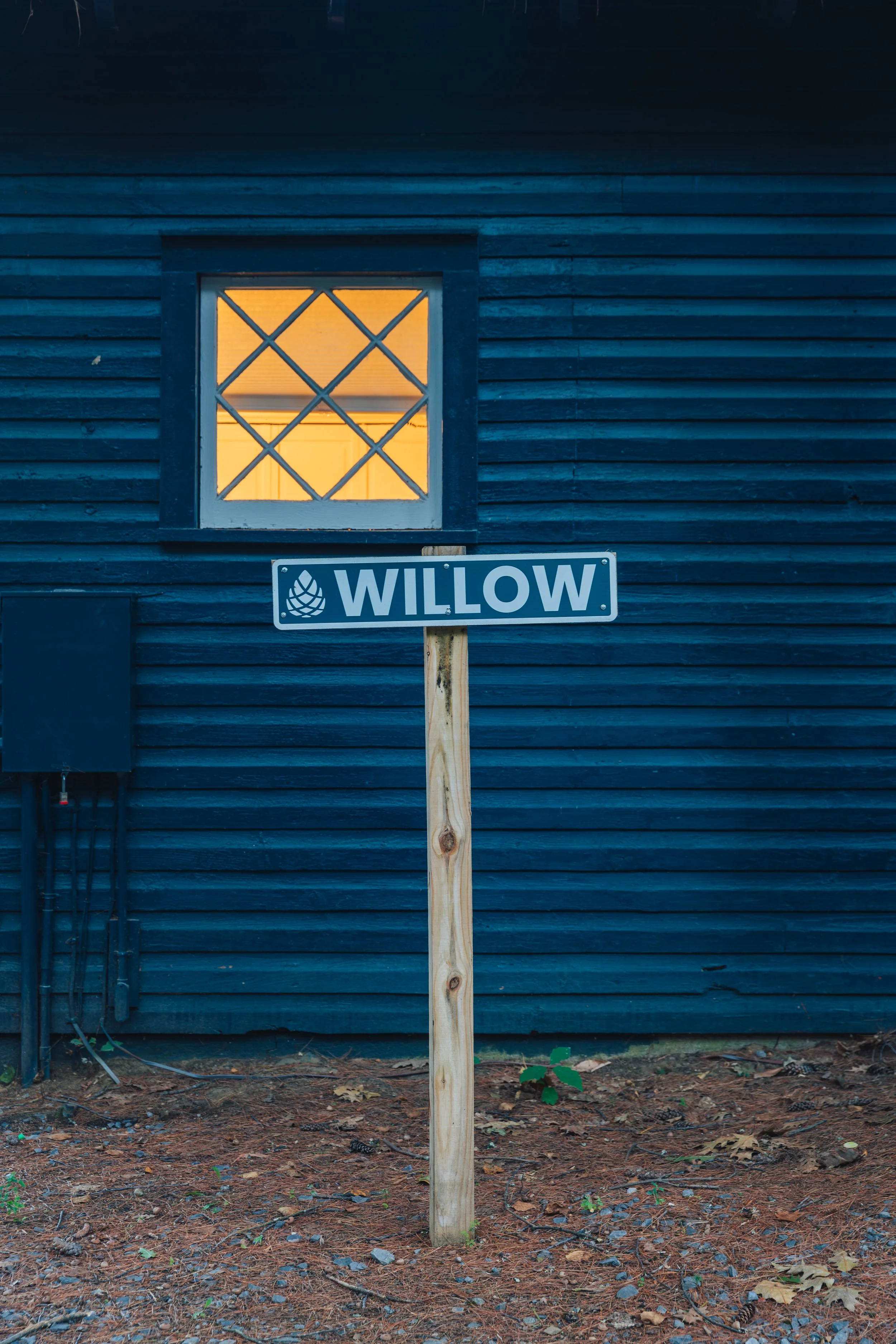 Welcome to the 'Willow' cabin,  a blue wooden house with a window that has a warm yellow light on inside.