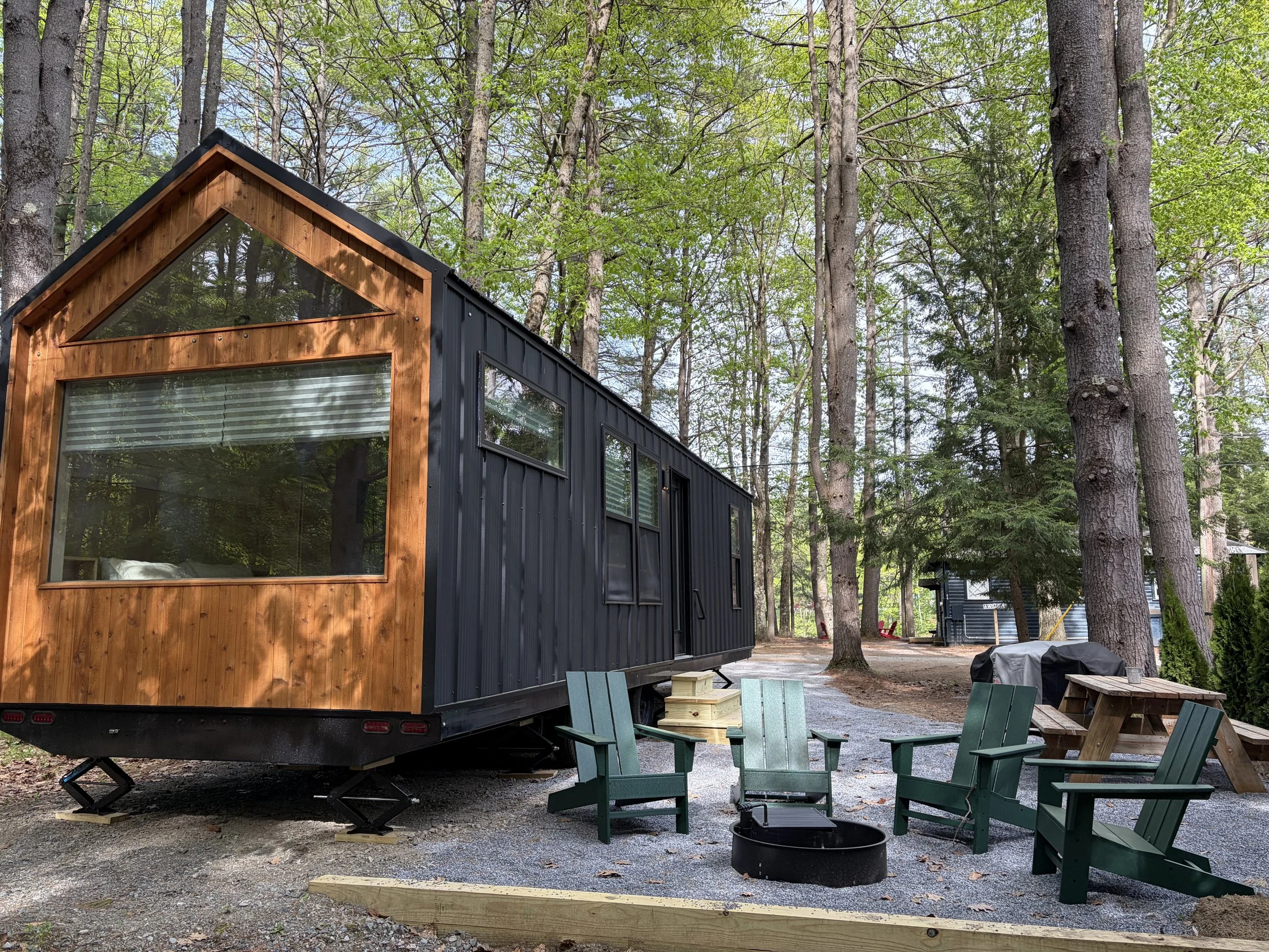 Escape to a tiny house with dark siding and a wooden framed window, situated in a forested area with tall trees and outdoor seating furniture.