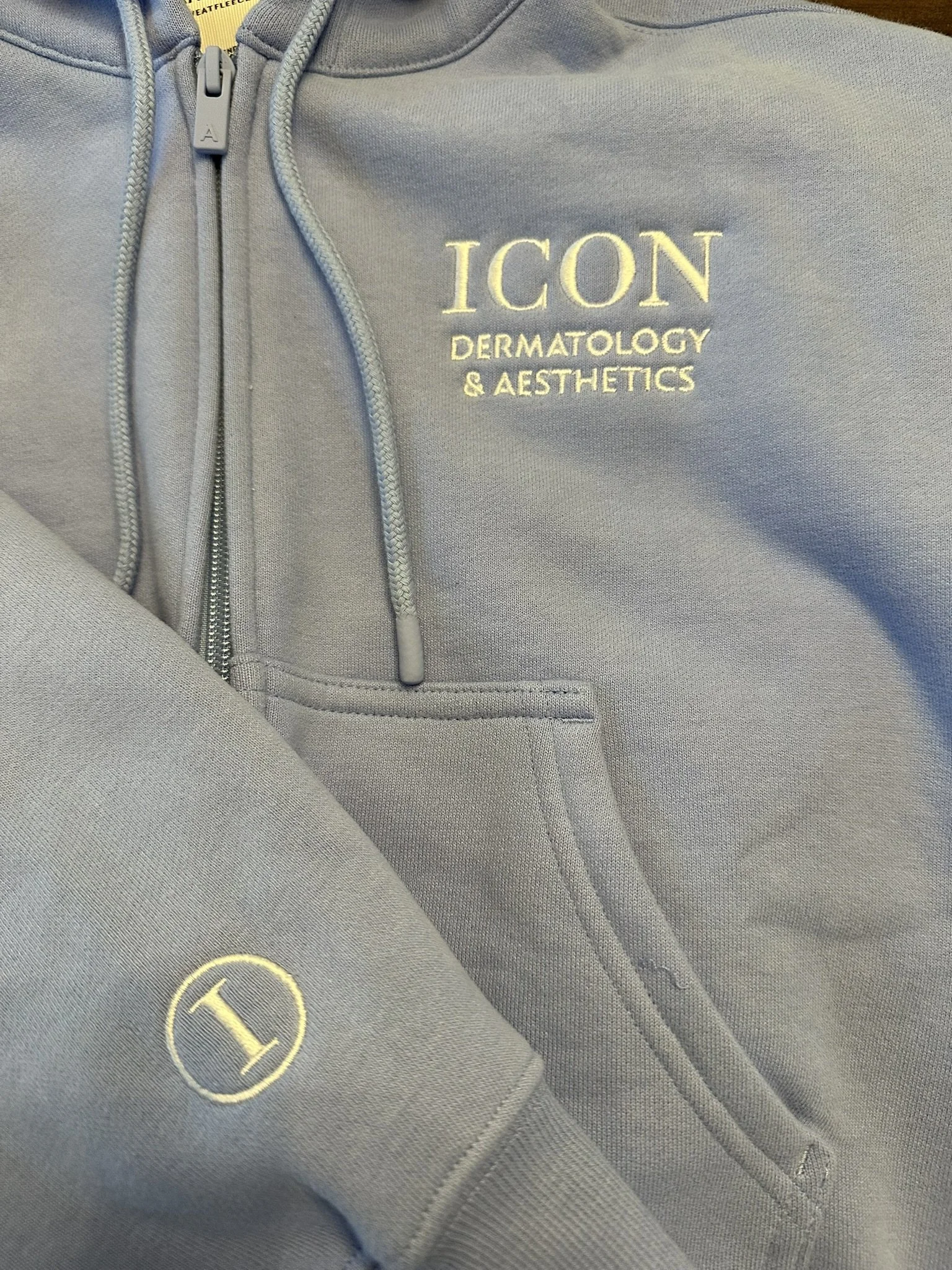 Gray hoodie with embroidered text reading "ICON Dermatology & Aesthetics" and a small circular logo with an "I" on the sleeve.