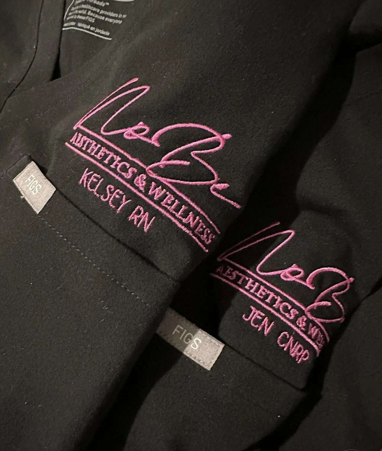 Two black scrubs with pink embroidery that says "Liez" and "Liez Athletics & Wellness," with names "Kelsey RN" and "Jen CNMP."