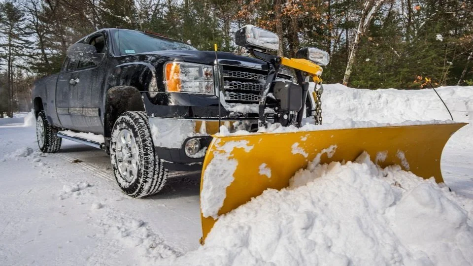 Black pickup truck with a yellow snow plow clearing snow on a snowy road, with trees in the background.