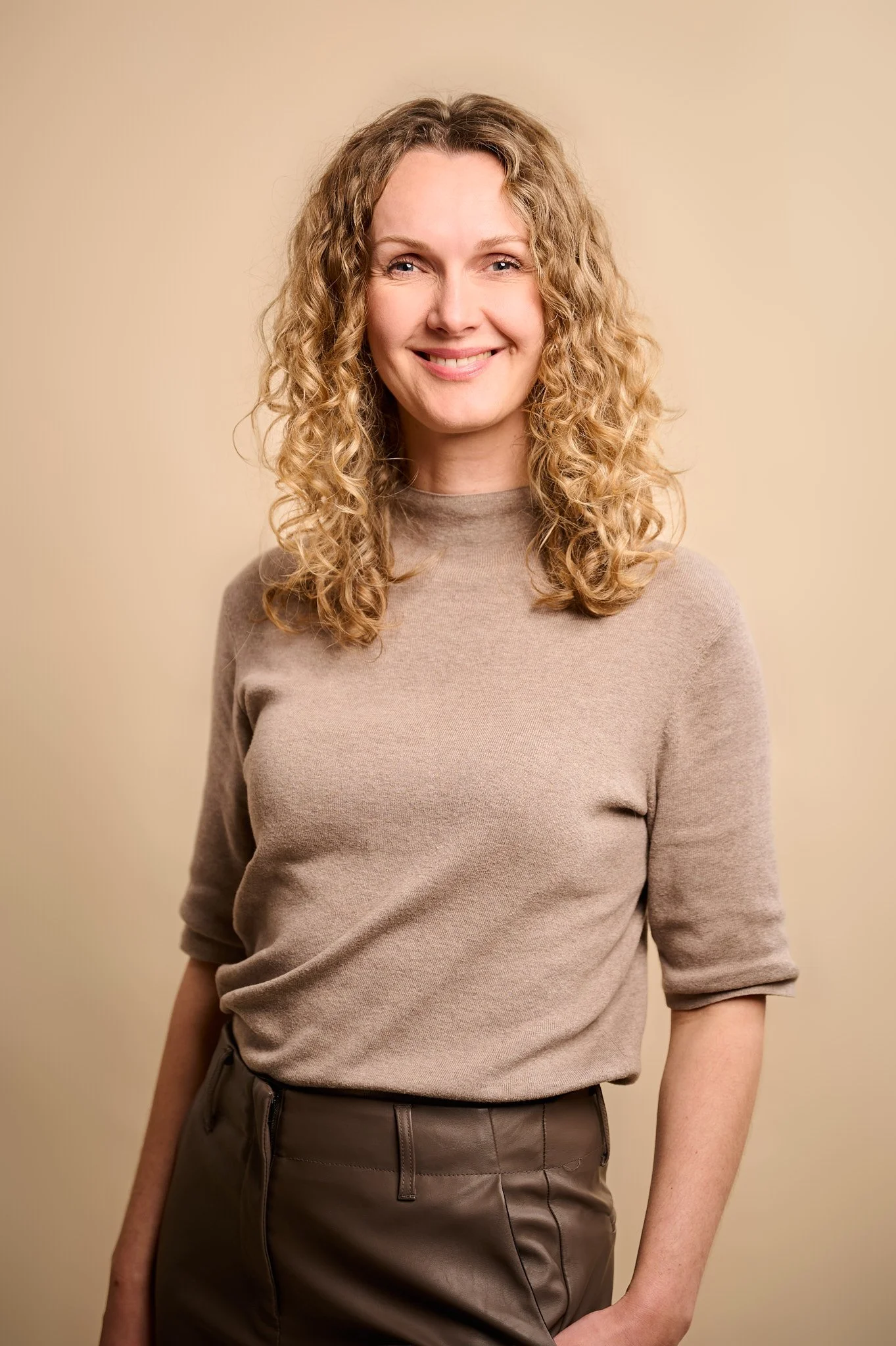 Portret van een vrouw met curly blond haar en blauwe ogen, gekleed in een beige trui, lachend tegen een effen beige achtergrond.