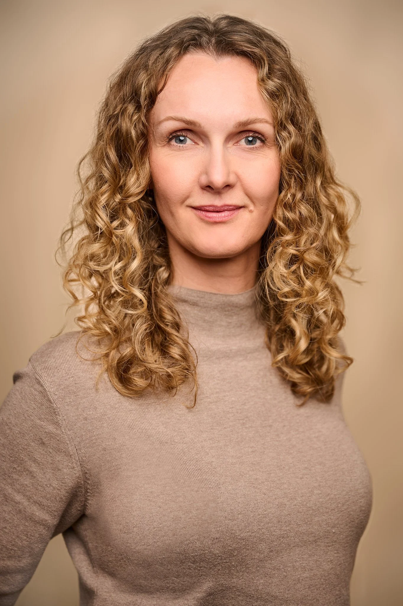 Portret van een vrouw met curly blond haar en blauwe ogen, gekleed in een beige trui, lachend tegen een effen beige achtergrond.