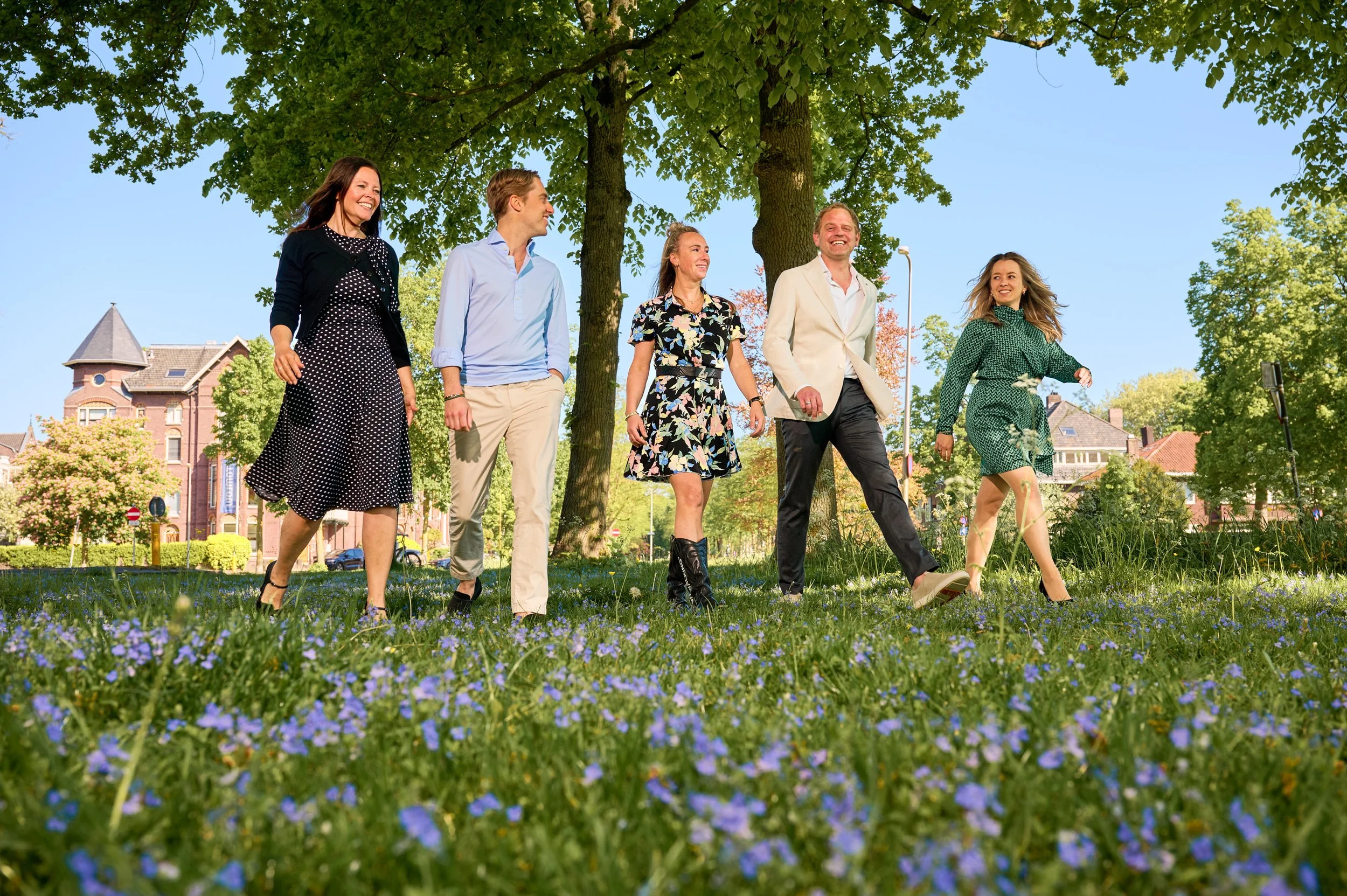 Vijf mensen wandelen lachend in een park onder een grote boom, met bloemen op de voorgrond en huizen op de achtergrond op een zonnige dag.