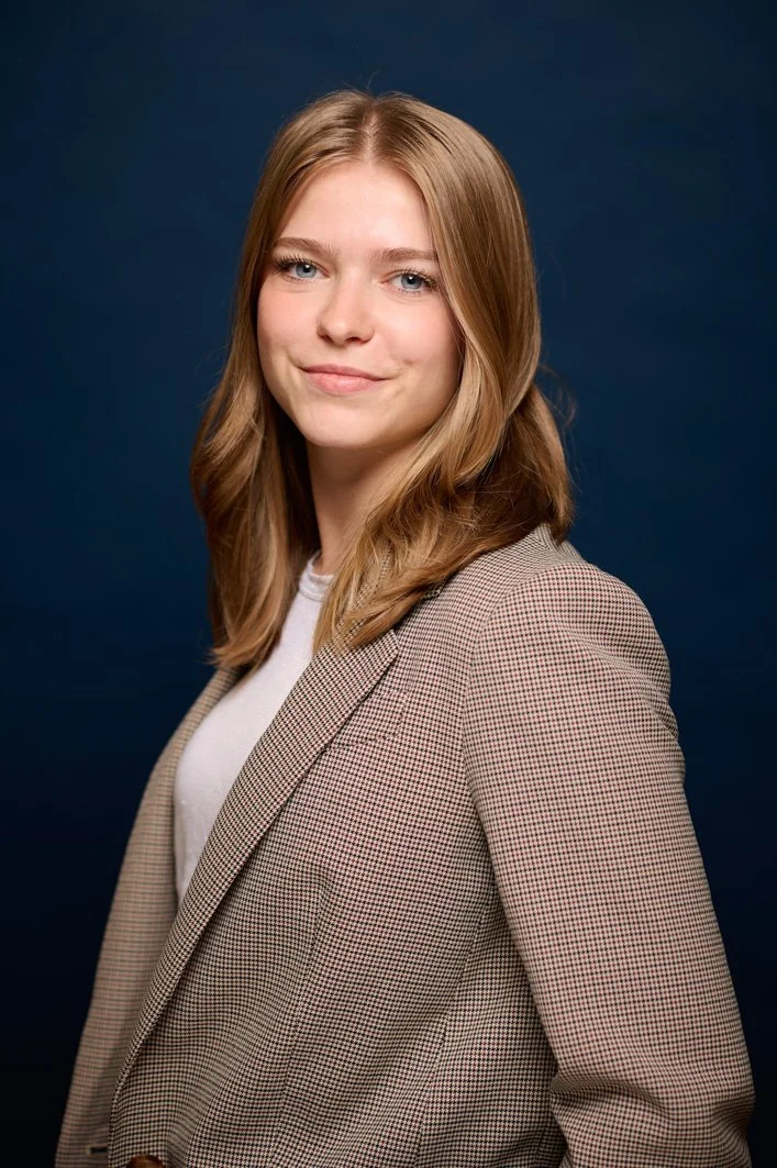 Portret van een jonge vrouw met bruin haar, gekleed in een beige geruite blazer en een witte top, lachend voor een donkere achtergrond.